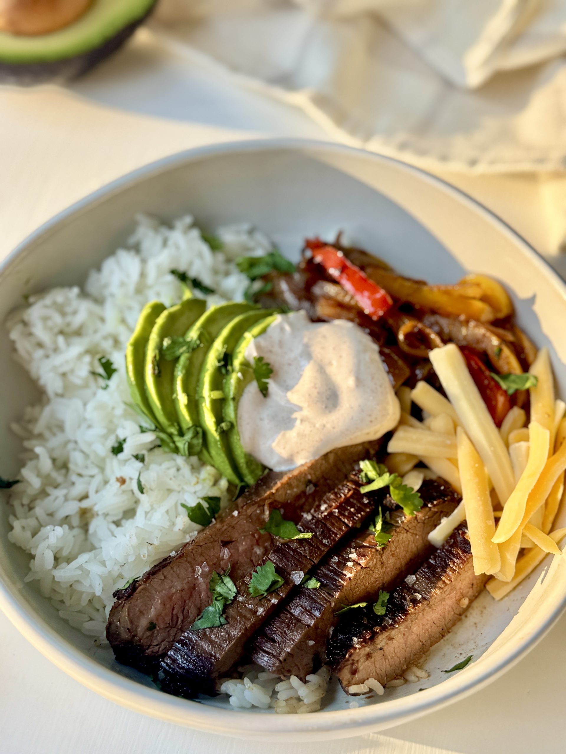 A bowl filled with white rice, sliced grilled steak, avocado slices, sautéed peppers and onions, shredded cheese, and a dollop of creamy sauce, garnished with chopped herbs.