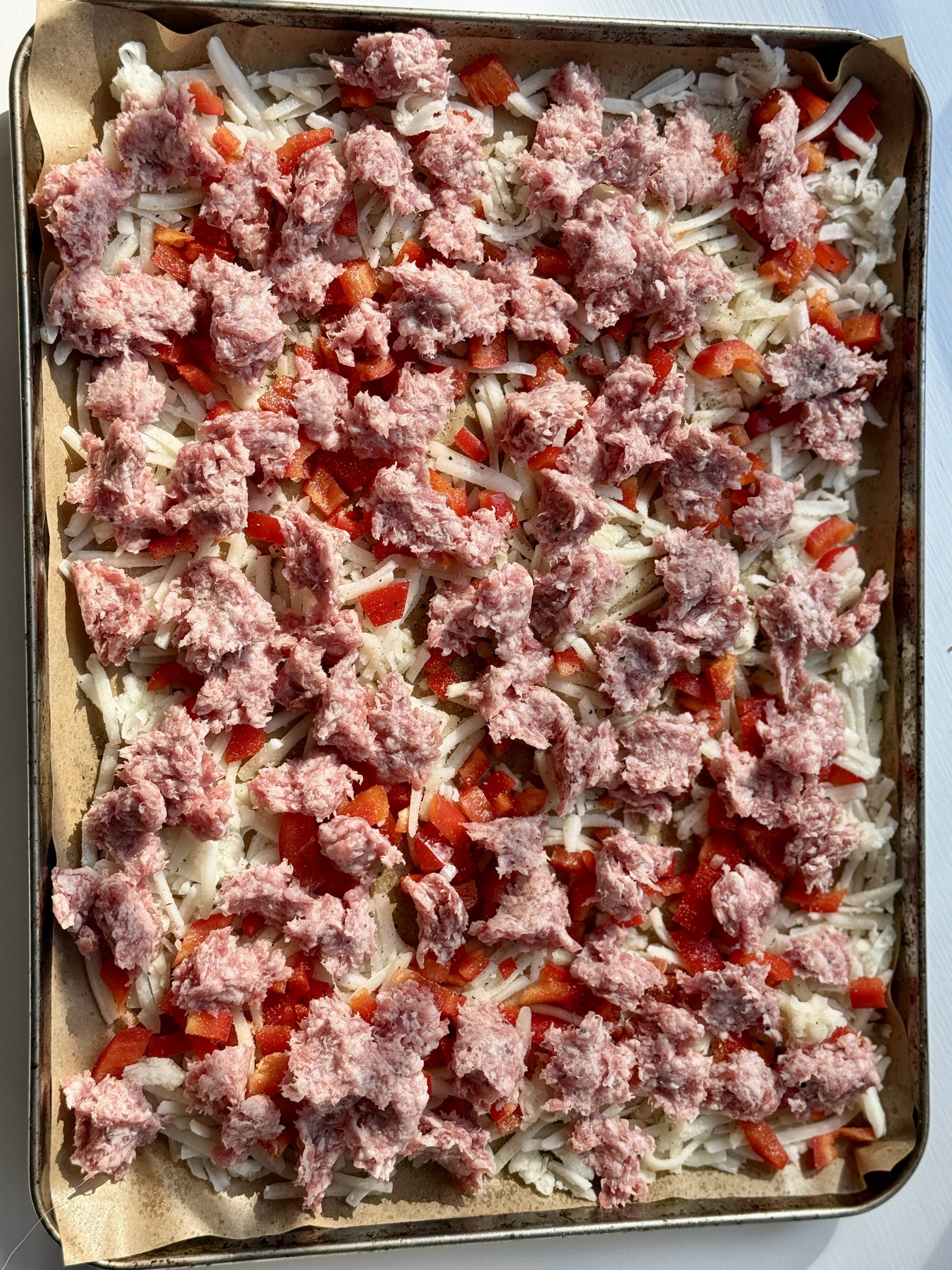 Raw breakfast hash on a baking sheet with shredded potatoes, diced red bell peppers, and crumbled sausage, ready for cooking. Bright and fresh.