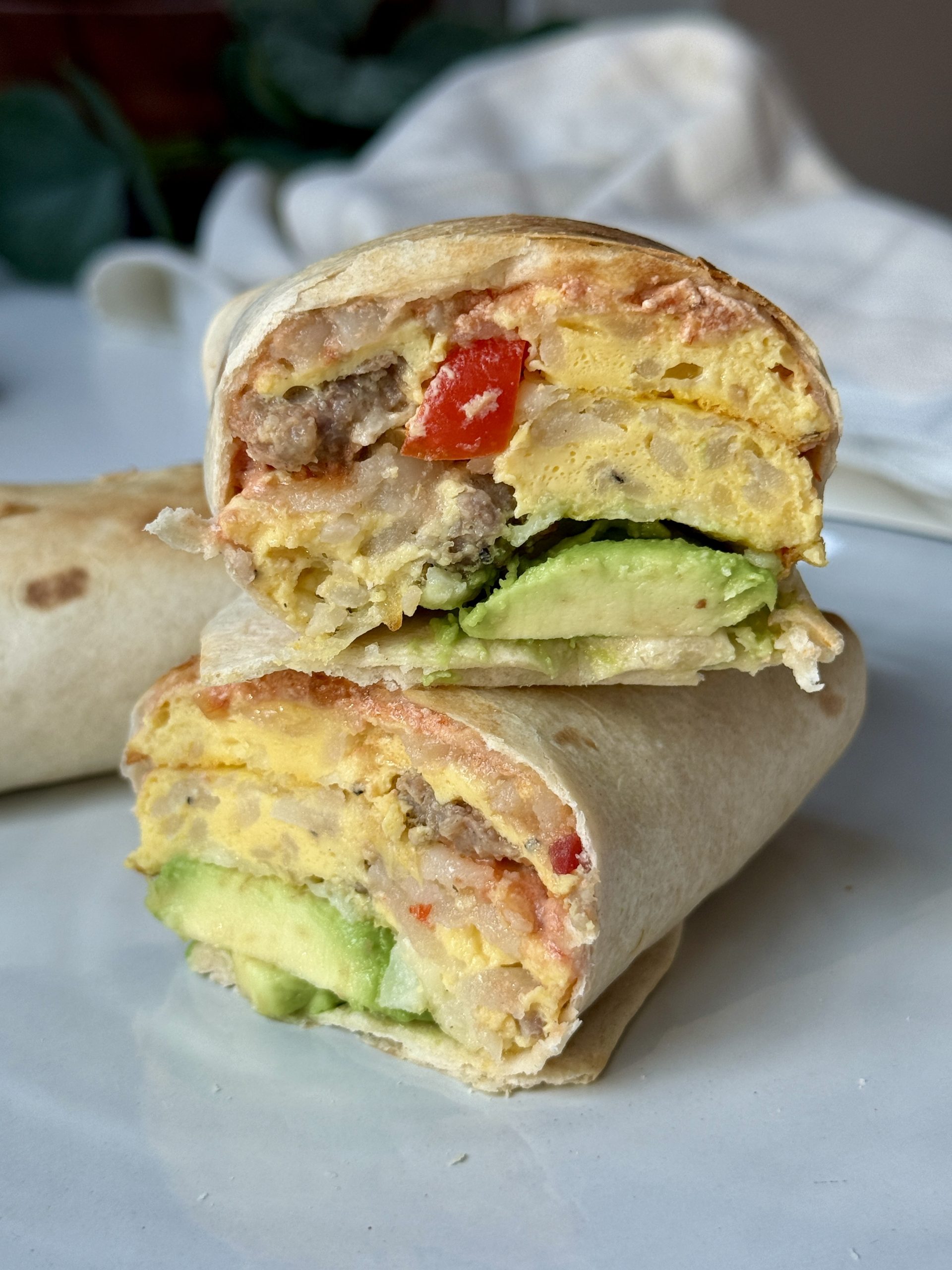 Close-up of a breakfast burrito cut in half, revealing layers of scrambled eggs, sausage, avocado, and red peppers. The setting is bright and inviting.