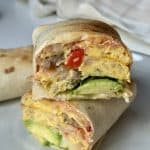 Close-up of a breakfast burrito cut in half, revealing layers of scrambled eggs, sausage, avocado, and red peppers. The setting is bright and inviting.