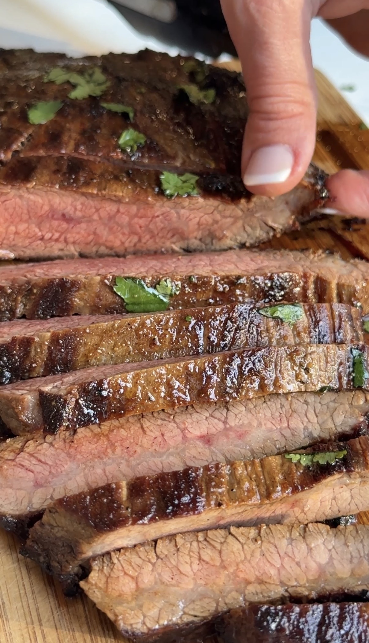 Close-up of juicy, grilled steak slices garnished with herbs on a wooden cutting board, with a hand holding one slice at the top of the image.