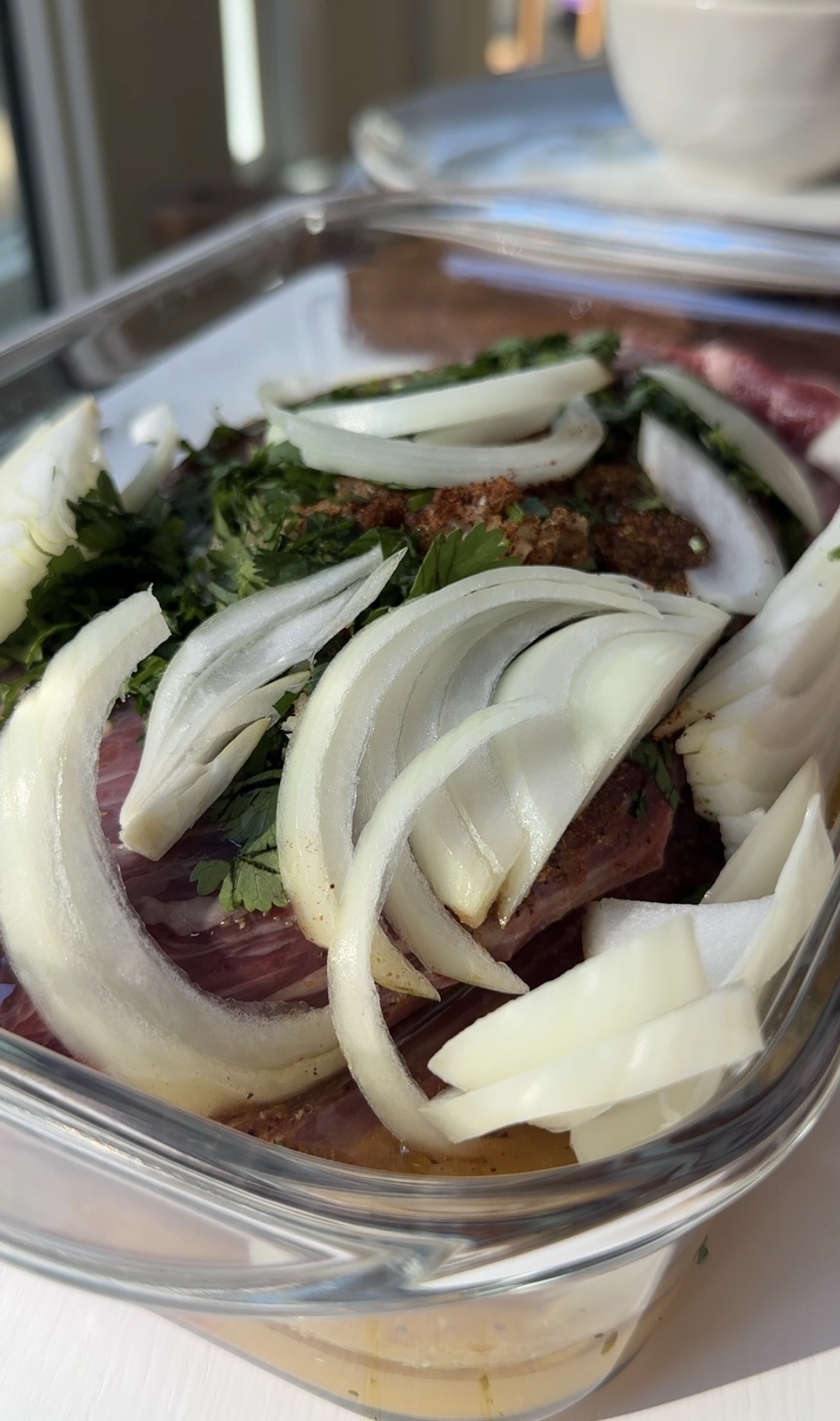Sliced onions and fresh herbs are layered over raw seasoned meat in a clear glass baking dish, ready for cooking.