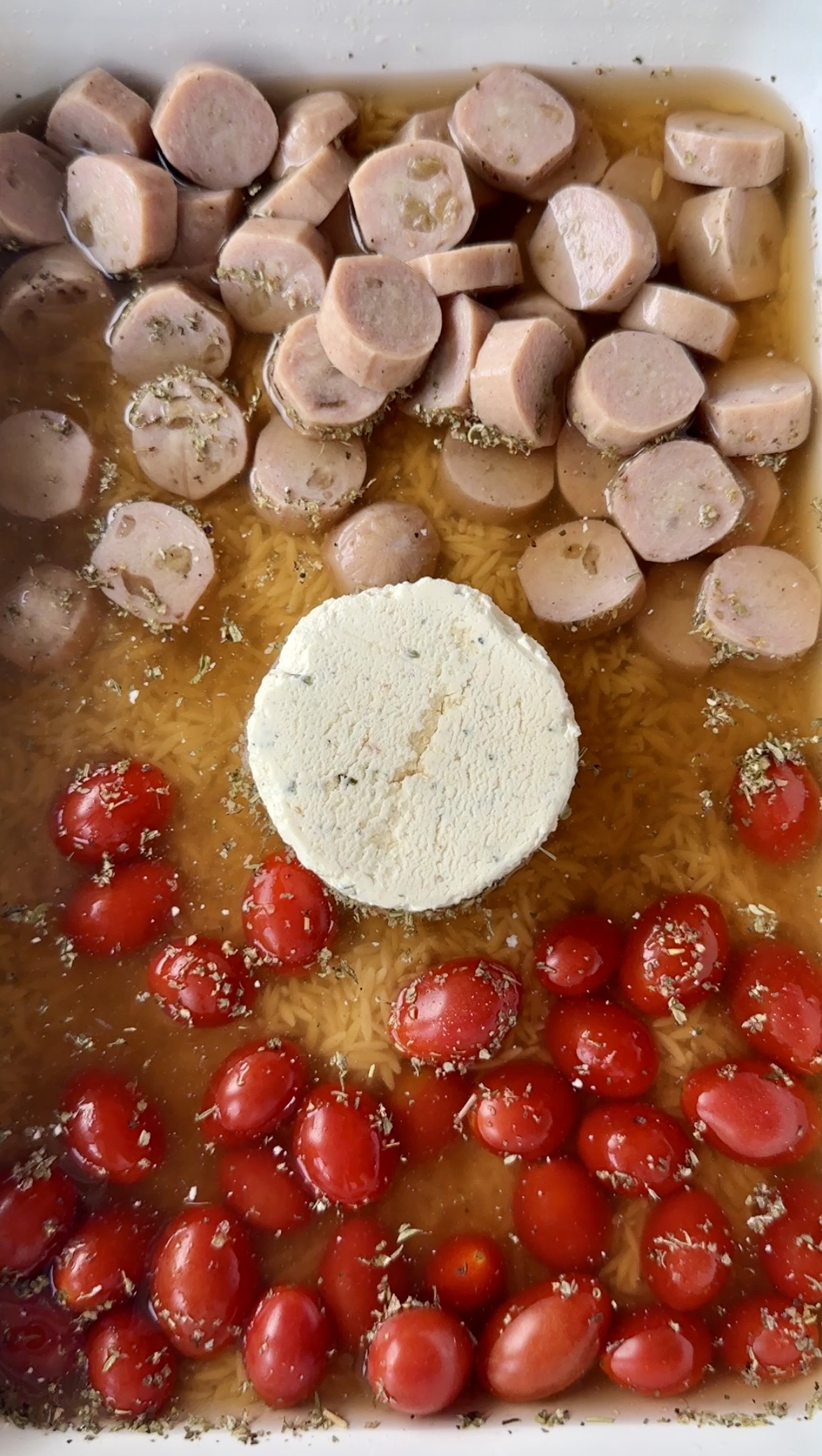 A baking dish filled with sliced sausages, grape tomatoes, a block of creamy cheese in the center, uncooked rice, and sprinkled herbs, all surrounded by broth.