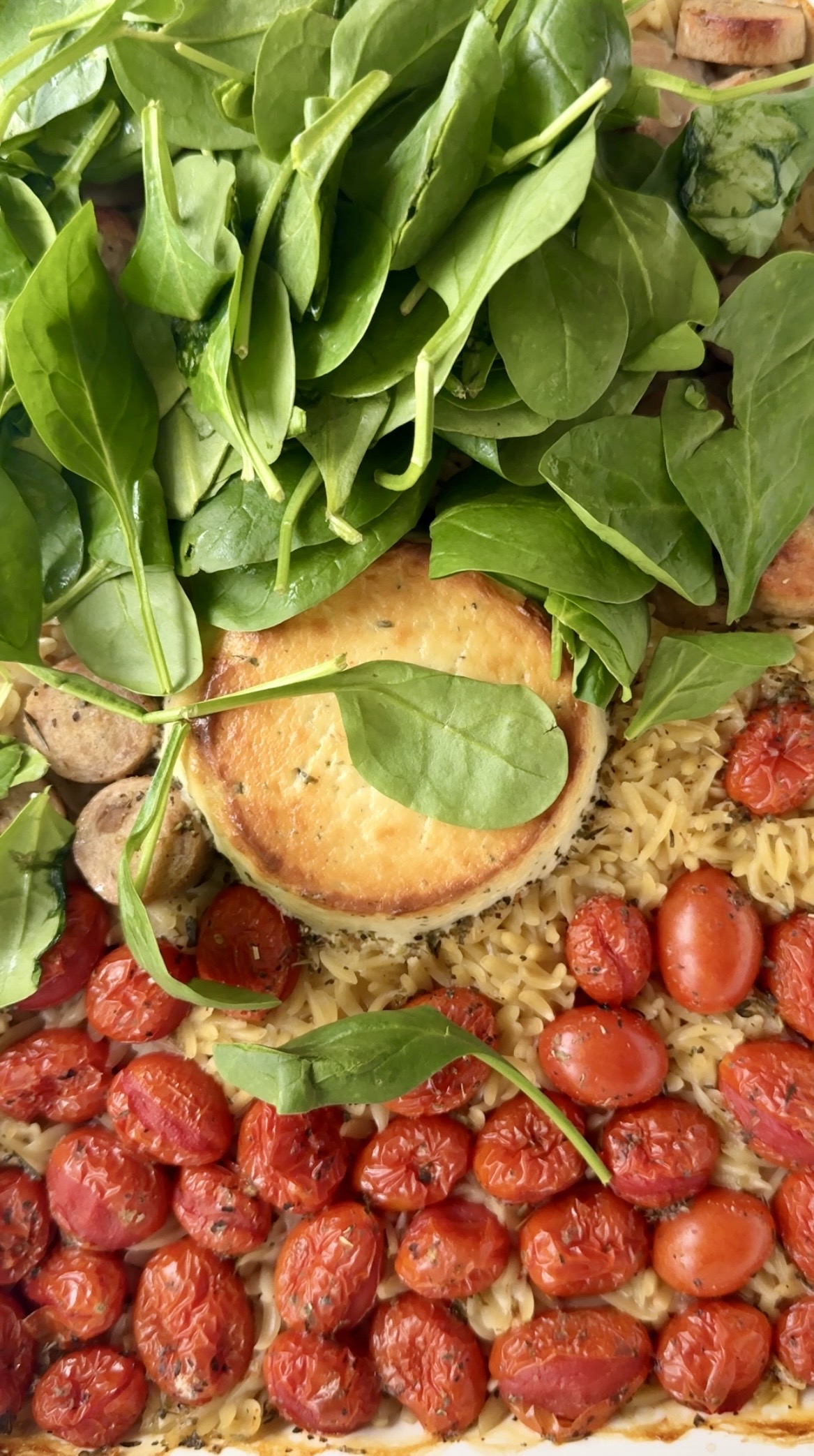 A tray filled with roasted cherry tomatoes, orzo pasta, mushrooms, a round block of baked cheese, and a generous handful of fresh spinach leaves on top.