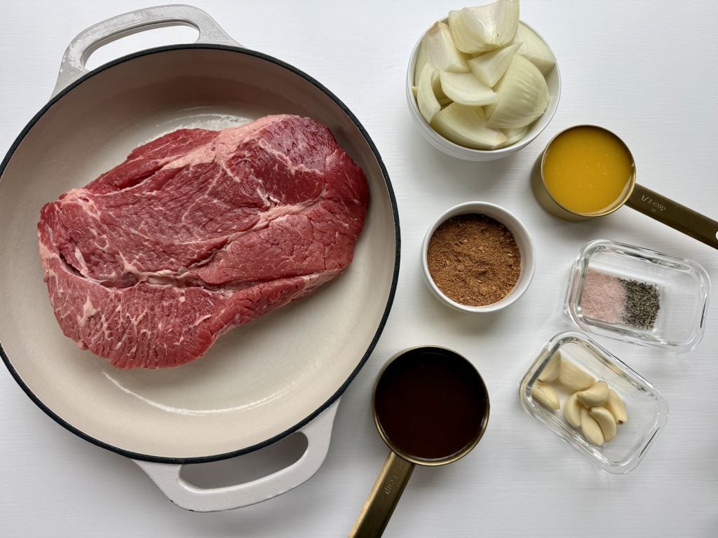 Overhead view of a white pot with a large raw beef roast, perfect for slow-braised chuck roast tacos, surrounded by bowls of cut onions, brown sugar, whole garlic cloves, salt and pepper, orange juice, and a cup of dark liquid on a white surface.