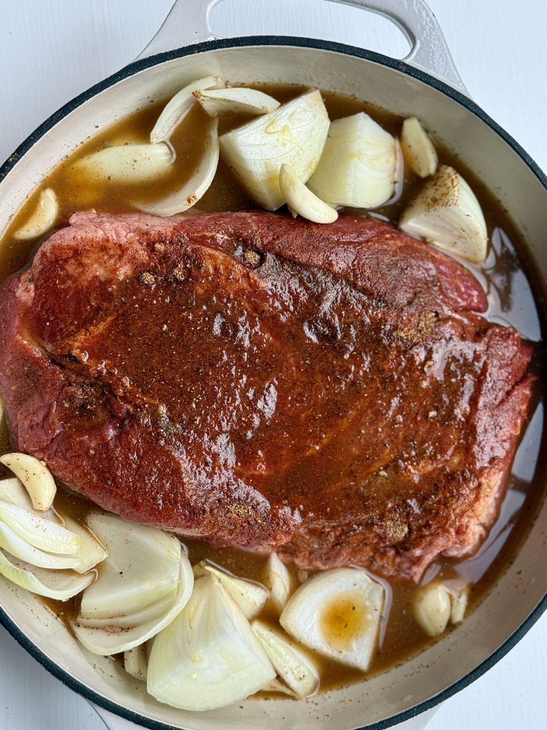 A raw seasoned beef roast, perfect for slow-braised chuck roast tacos, sits in a Dutch oven surrounded by chopped onions, whole garlic cloves, and broth, ready to be cooked.