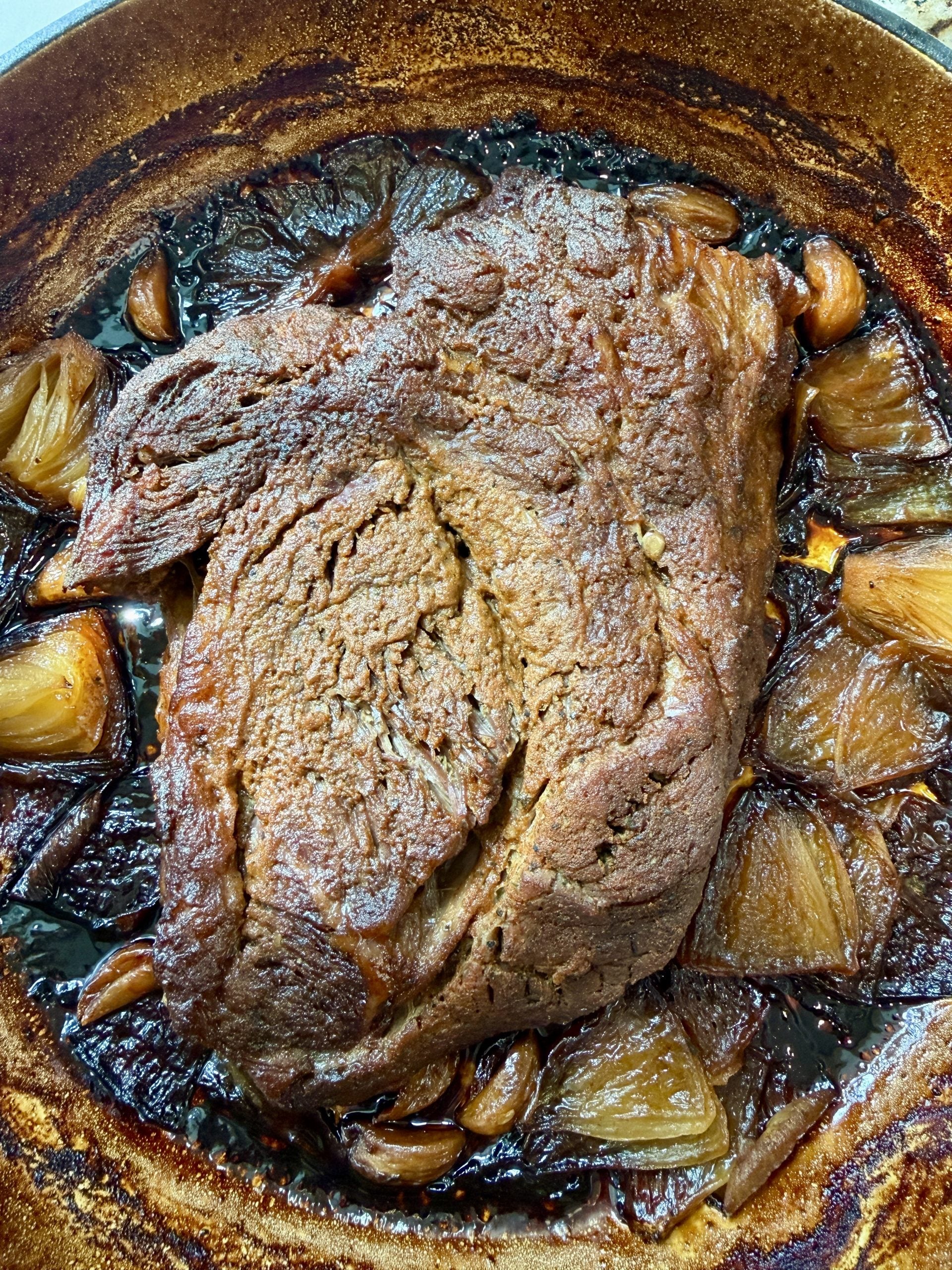 A slow-braised beef roast sits in a brown roasting pan surrounded by caramelized onions and whole garlic cloves, with crispy edges and a deeply browned exterior.