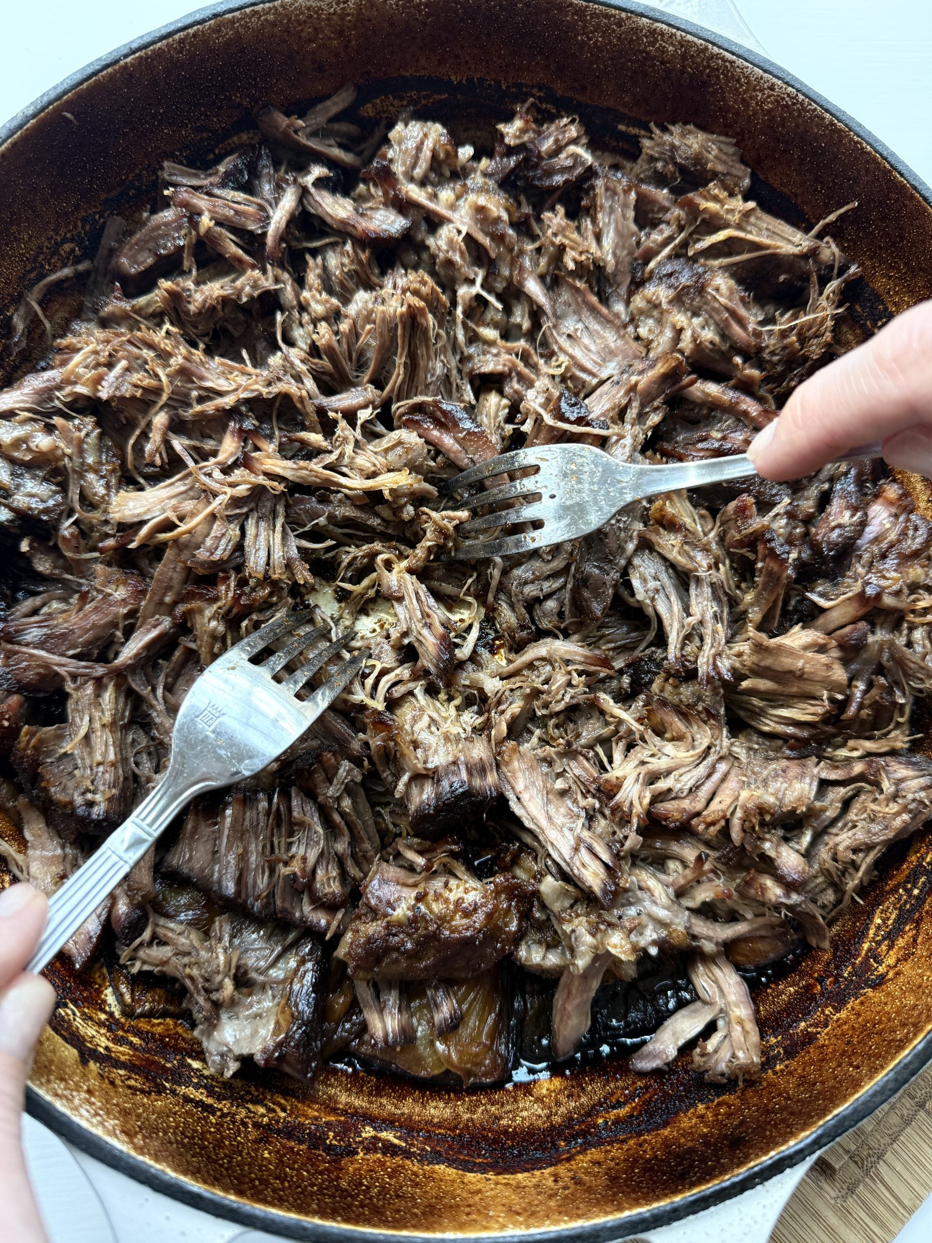 Two hands use forks to shred tender, slow-braised beef in a large, dark-rimmed skillet. The meat, browned and moist with taco seasoning, fills the pan—perfect for flavorful chuck roast tacos.