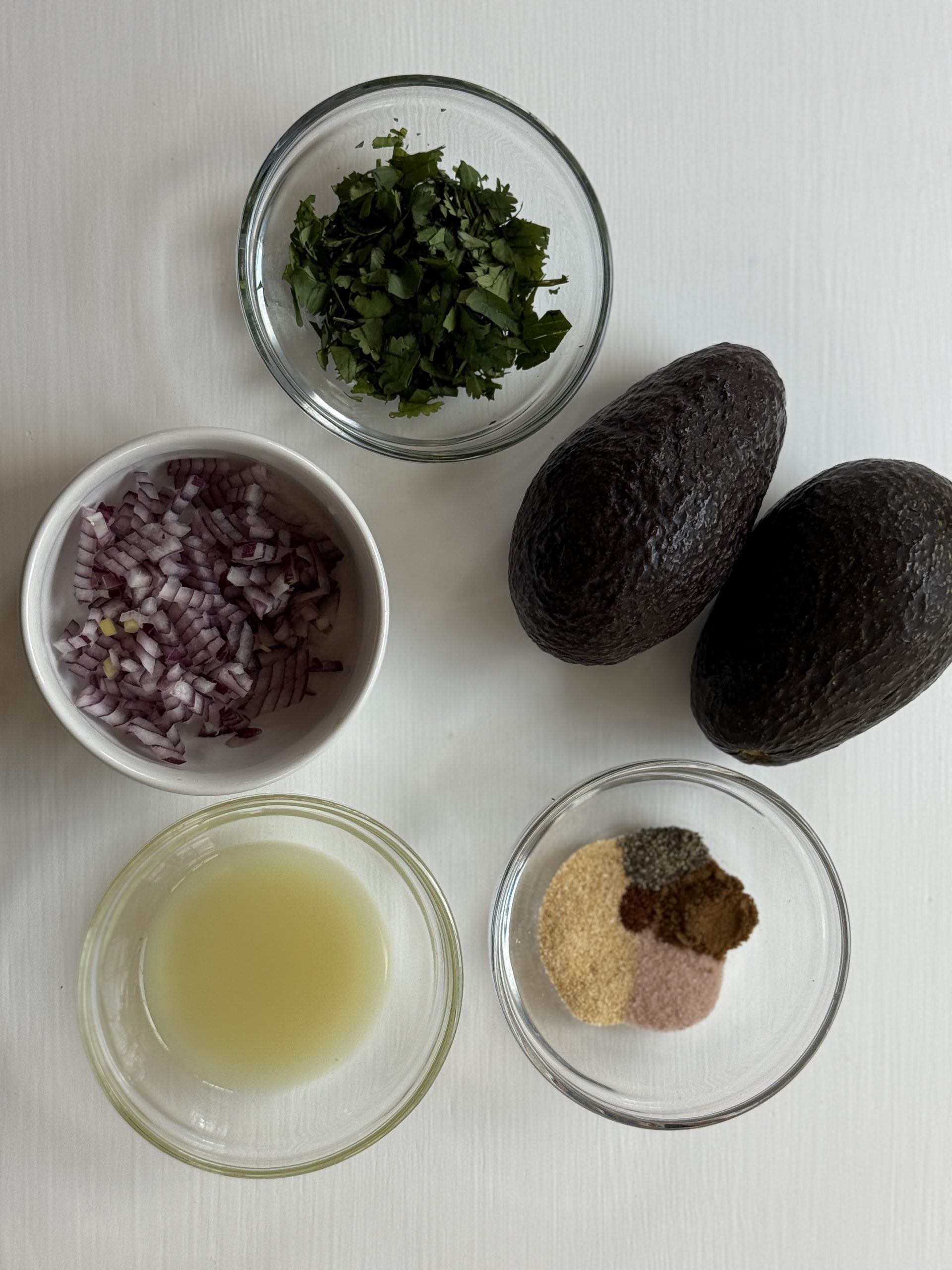 Five ingredients for guacamole on a white surface: chopped cilantro, two whole avocados, diced red onion, a bowl of lime juice, and a bowl with various spices.