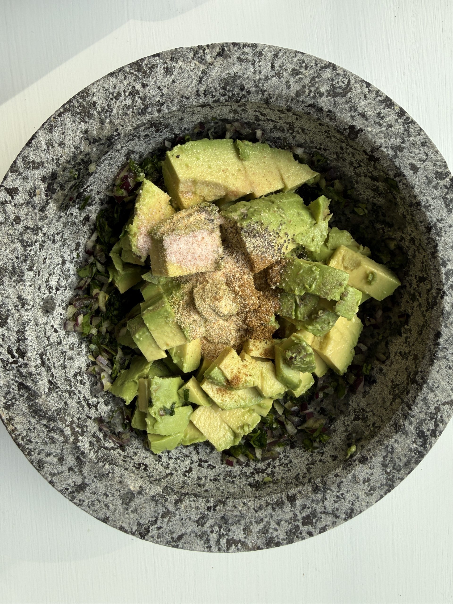 A stone bowl filled with chopped avocado, minced red onion, and seasonings, ready to be mixed, sits on a light-colored surface.