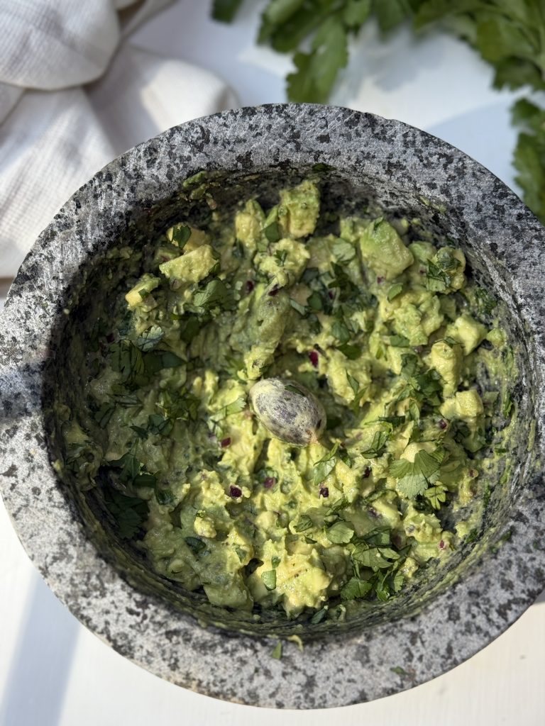 A stone mortar filled with freshly made guacamole, topped with chopped cilantro and a single avocado pit in the center. Parsley leaves and a white cloth are visible in the background.