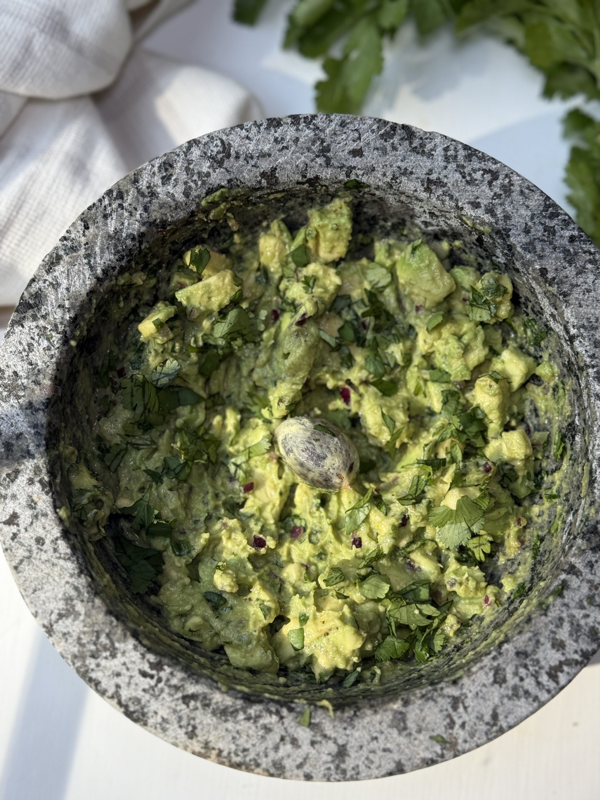 A stone mortar filled with freshly made guacamole, topped with chopped cilantro and a single avocado pit in the center. Parsley leaves and a white cloth are visible in the background.