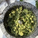 A stone mortar filled with chunky guacamole, garnished with chopped cilantro, sits on a light surface with a pestle inside. Fresh cilantro leaves and a white cloth are in the background.
