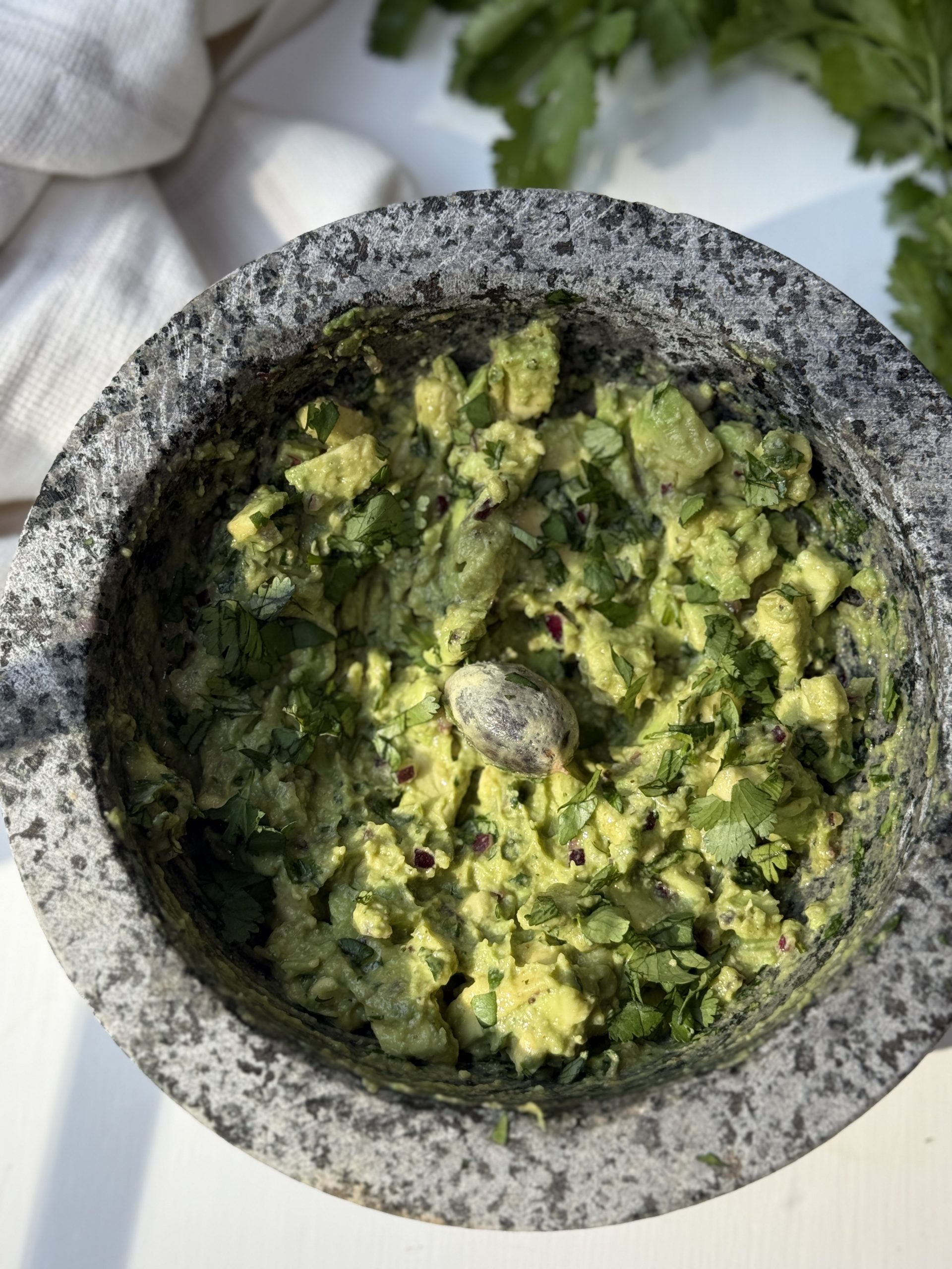 A stone mortar filled with chunky guacamole, garnished with chopped cilantro, sits on a light surface with a pestle inside. Fresh cilantro leaves and a white cloth are in the background.