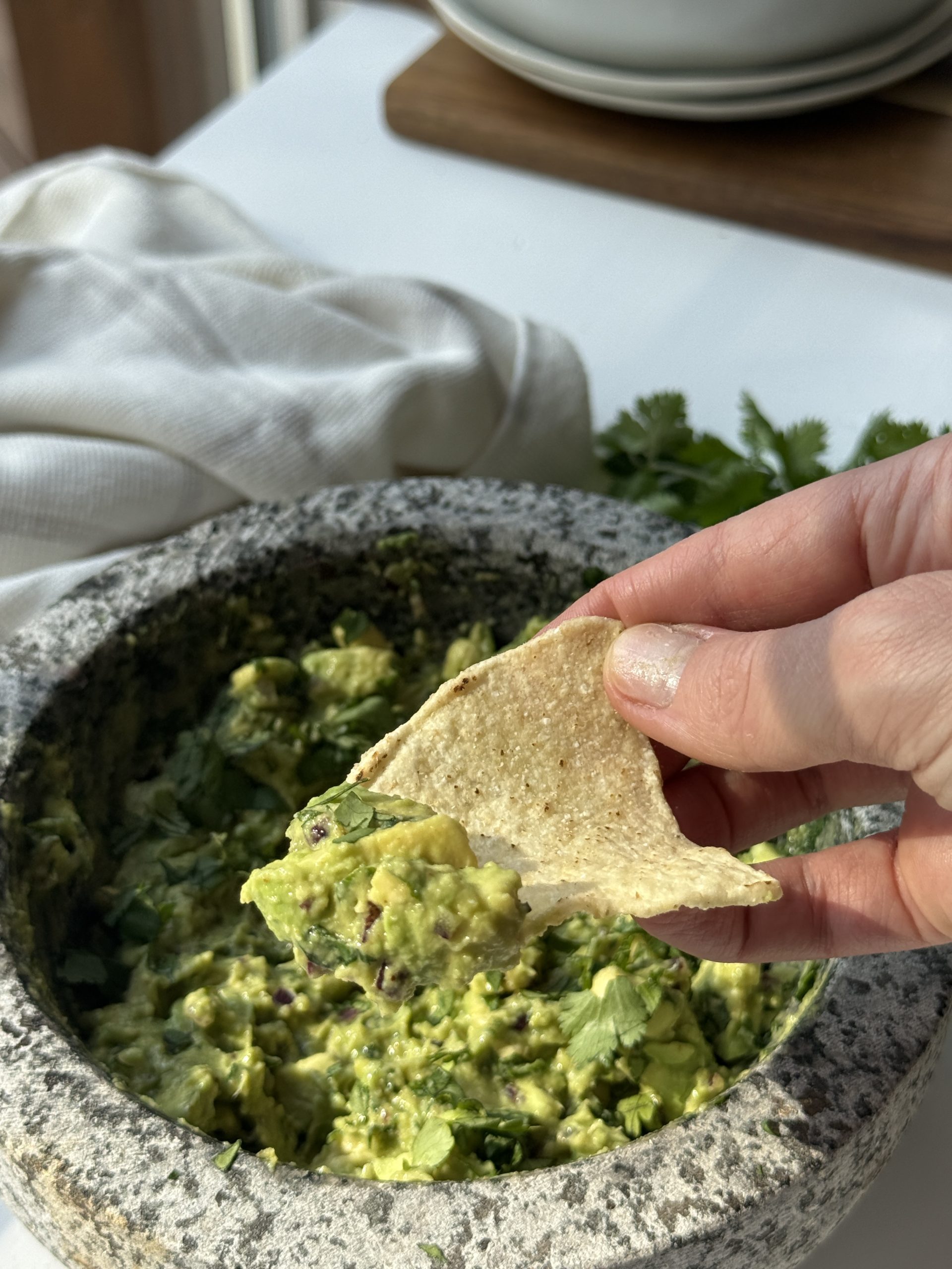 A hand holds a tortilla chip above a stone bowl filled with chunky guacamole, garnished with fresh cilantro. A white cloth and plates are in the background.