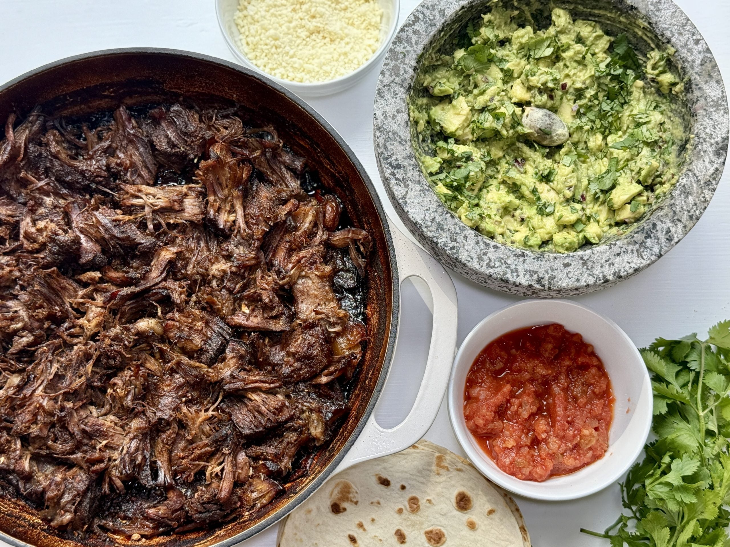 A skillet of slow-braised shredded beef with crispy edges, a stone bowl of guacamole, salsa, cheese, fresh cilantro, and a flour tortilla on a white surface—perfectly arranged for chuck roast tacos.