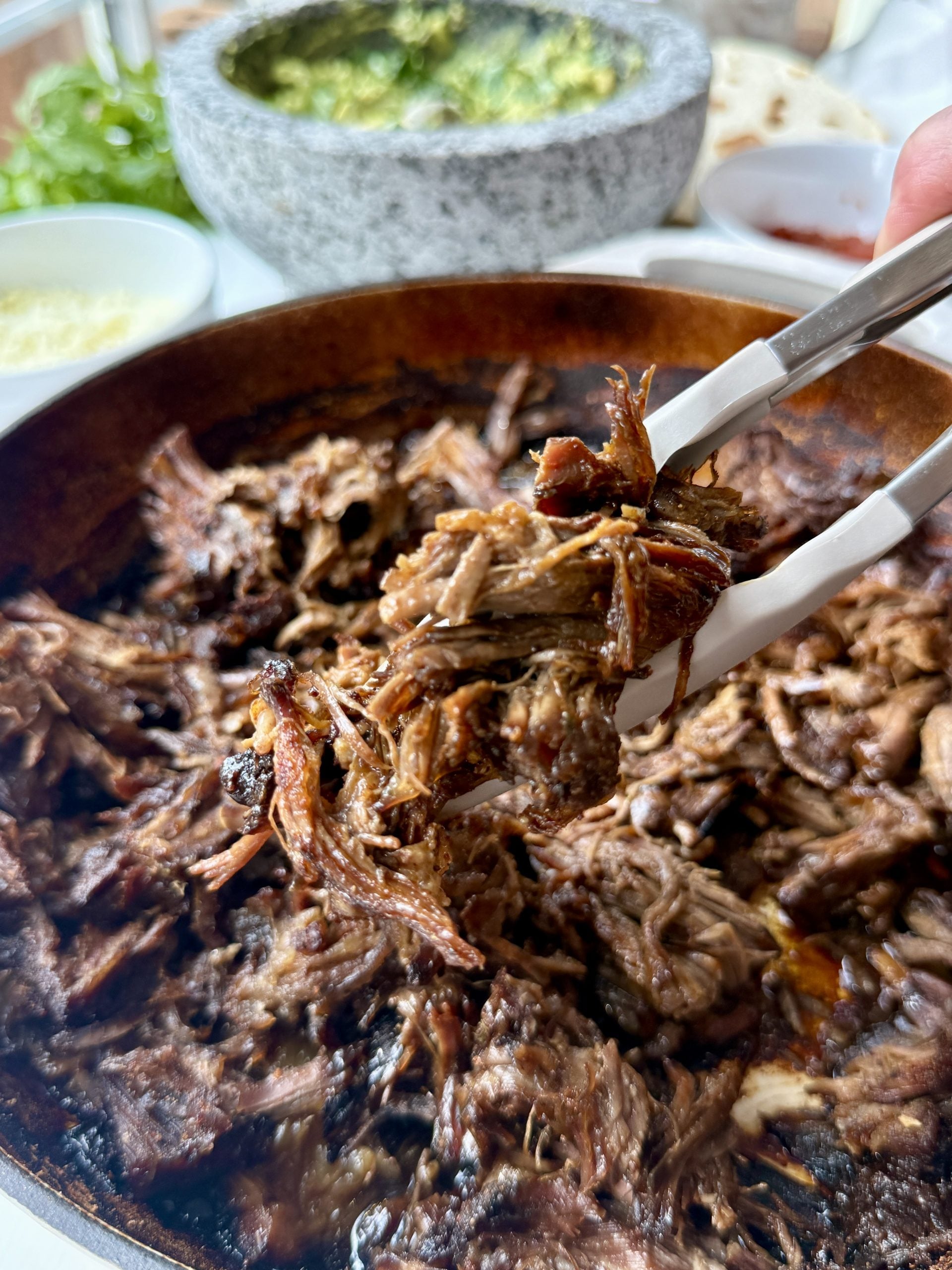 Slow-braised, shredded beef for chuck roast tacos is lifted with tongs from a skillet, with bowls of guacamole, cheese, and tortillas blurred in the background—perfect after hours in the oven.