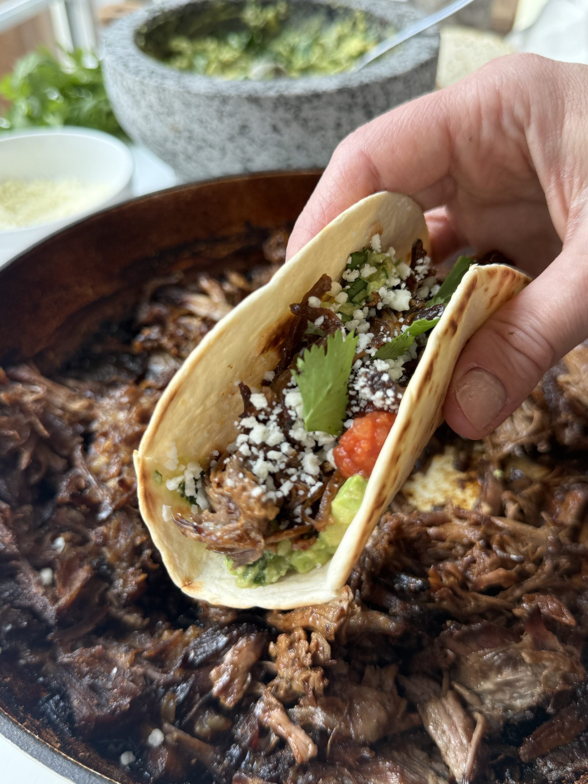 A hand holds a soft taco filled with slow-braised beef, cilantro, cheese, and salsa, with a skillet of chuck roast tacos and a bowl of green sauce in the background.