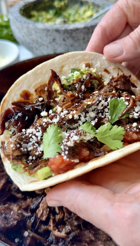 A hand holds a soft taco filled with slow-braised chuck roast tacos, garnished with crumbled cheese, cilantro, salsa, and guacamole. A bowl of green sauce is visible in the blurred background, hinting at flavorful taco seasoning.