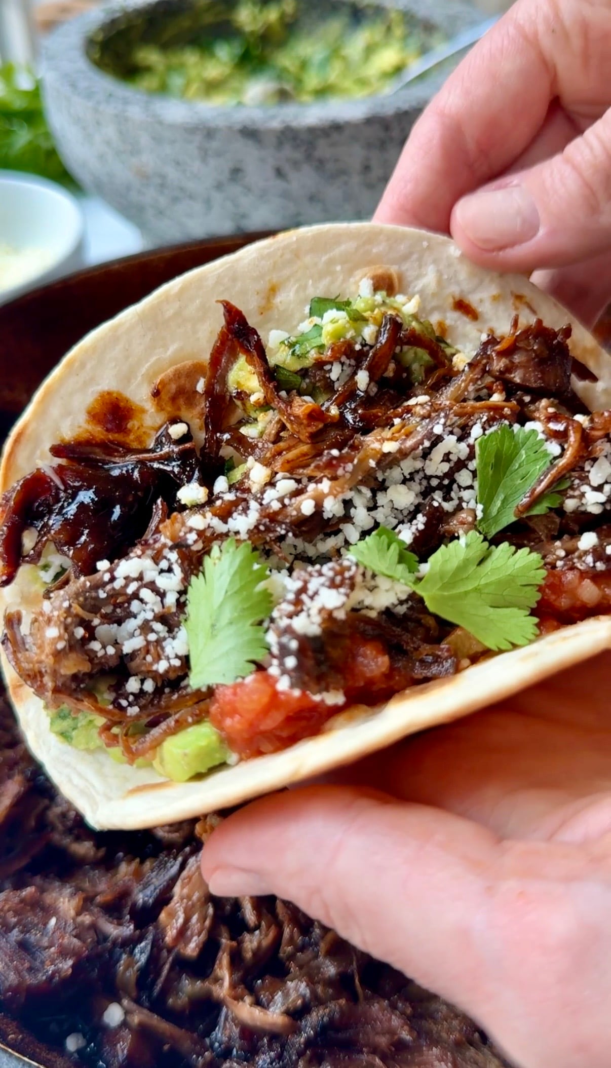 A close-up of a hand holding a soft taco filled with slow-braised chuck roast, salsa, guacamole, fresh cilantro leaves, and crumbled cheese, its crispy edges inviting against a blurred background showcasing more food.