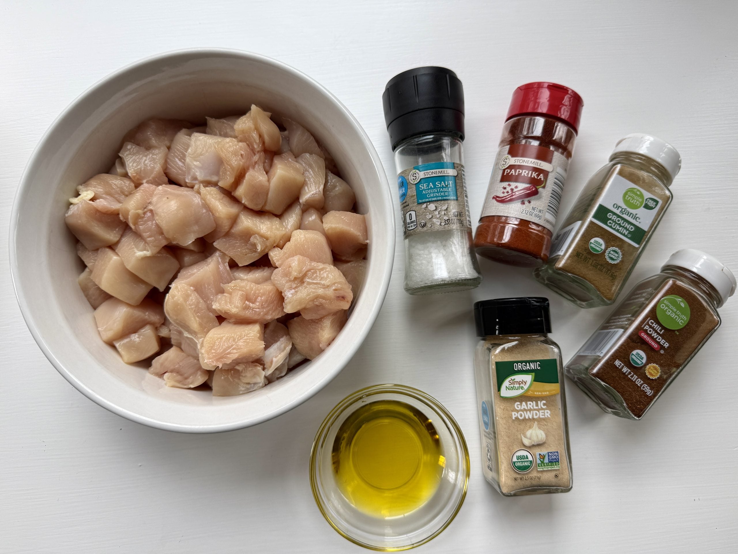 Raw cubed chicken in a white bowl surrounded by sea salt, paprika, cumin, coriander, garlic powder, and a small bowl of olive oil on a white surface.