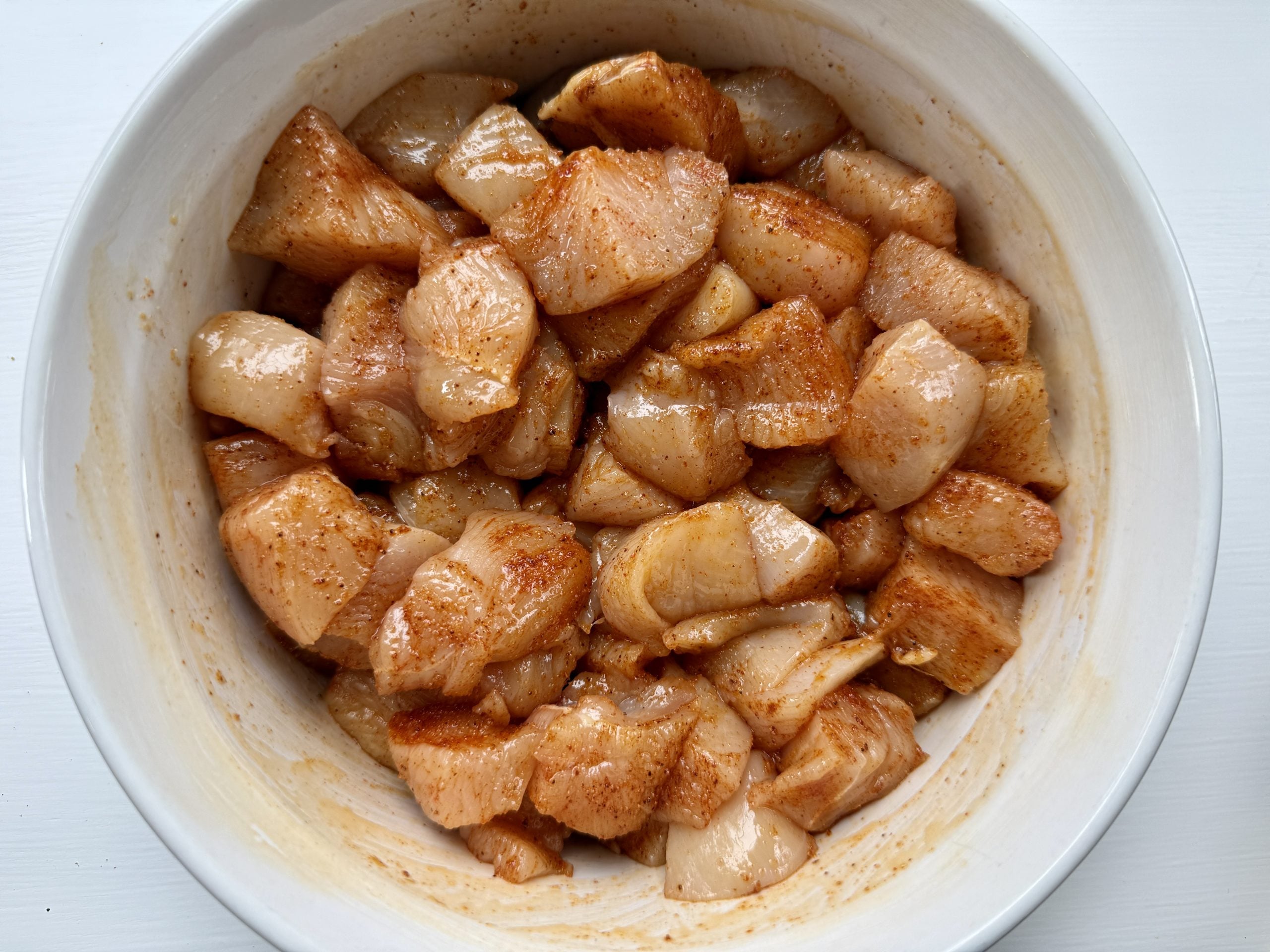 Diced raw chicken pieces coated in spices are placed in a white bowl. The seasoned meat has a glossy appearance, suggesting it is marinated.