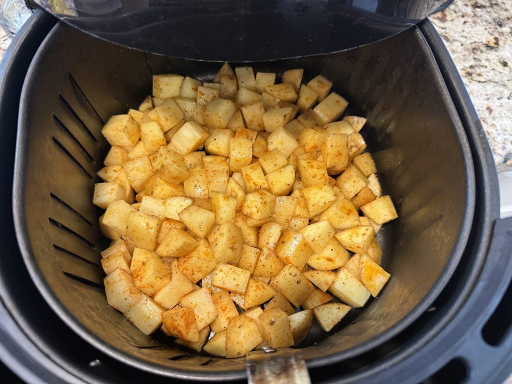 Diced potatoes seasoned with spices are evenly spread in an air fryer basket, ready for cooking. The scene conveys a sense of homemade preparation.