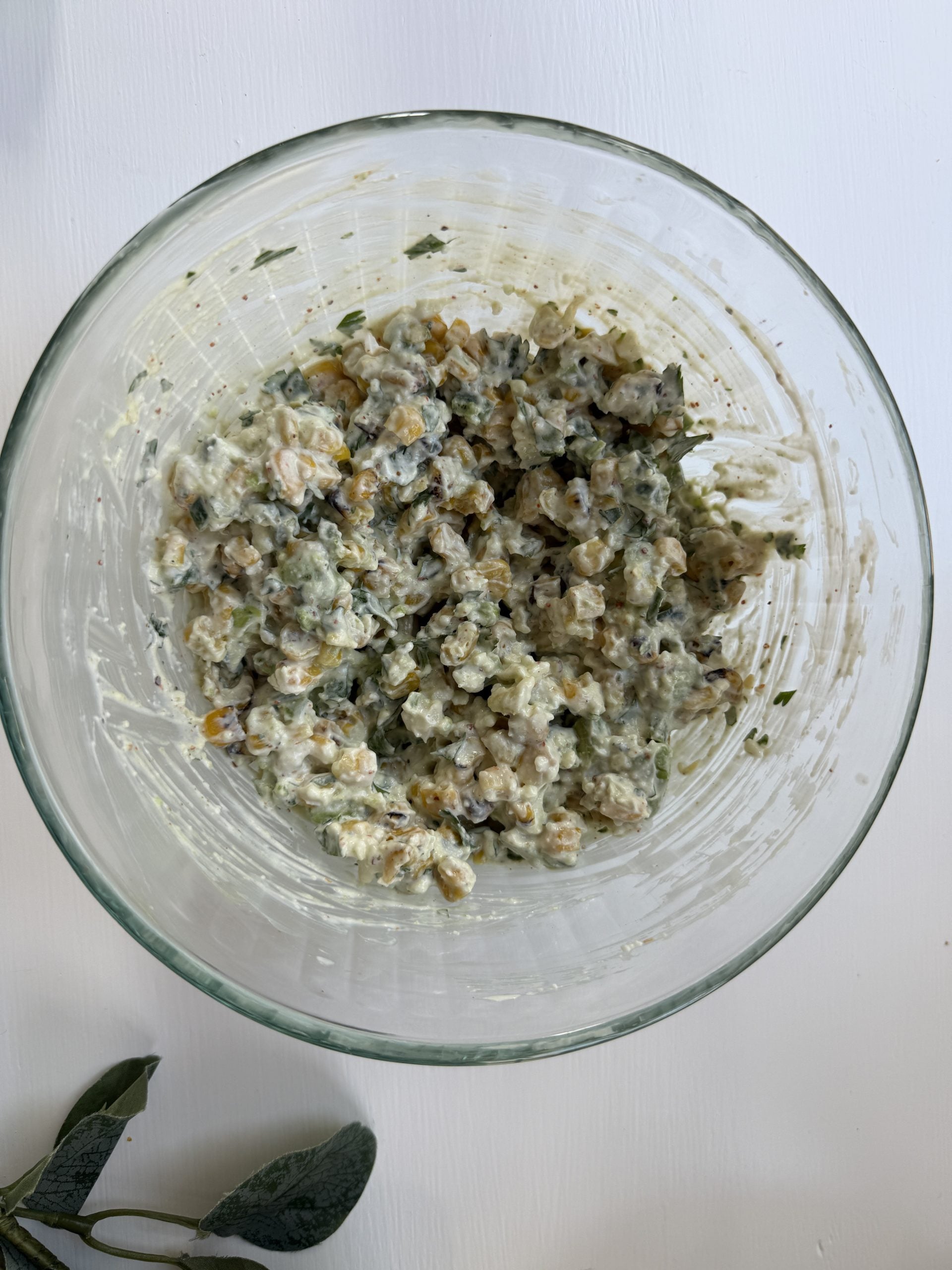 A glass bowl filled with creamy chickpea salad, featuring chopped herbs and visible green and orange vegetable bits. A fresh sage leaf is nearby on a white table.