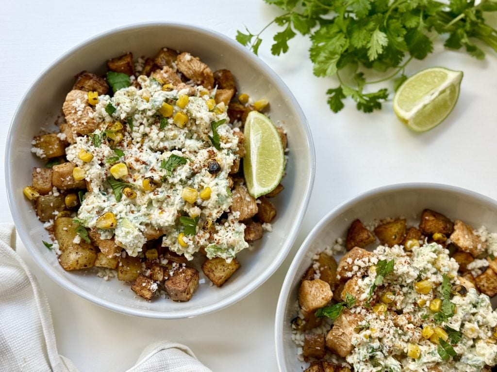 Two bowls filled with roasted potatoes, grilled chicken, and corn topped with creamy sauce and crumbled cheese. Garnished with lime and cilantro.