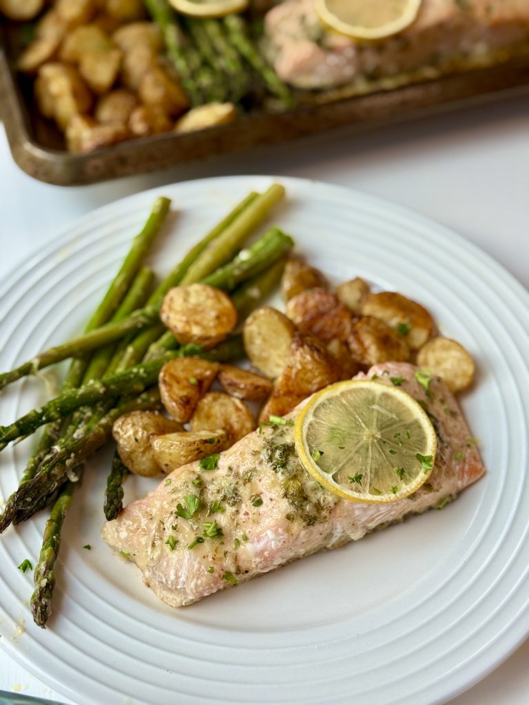 A plate on the table with salmon piece, topped with lemon, served with a side of roasted potatoes and asparagus 