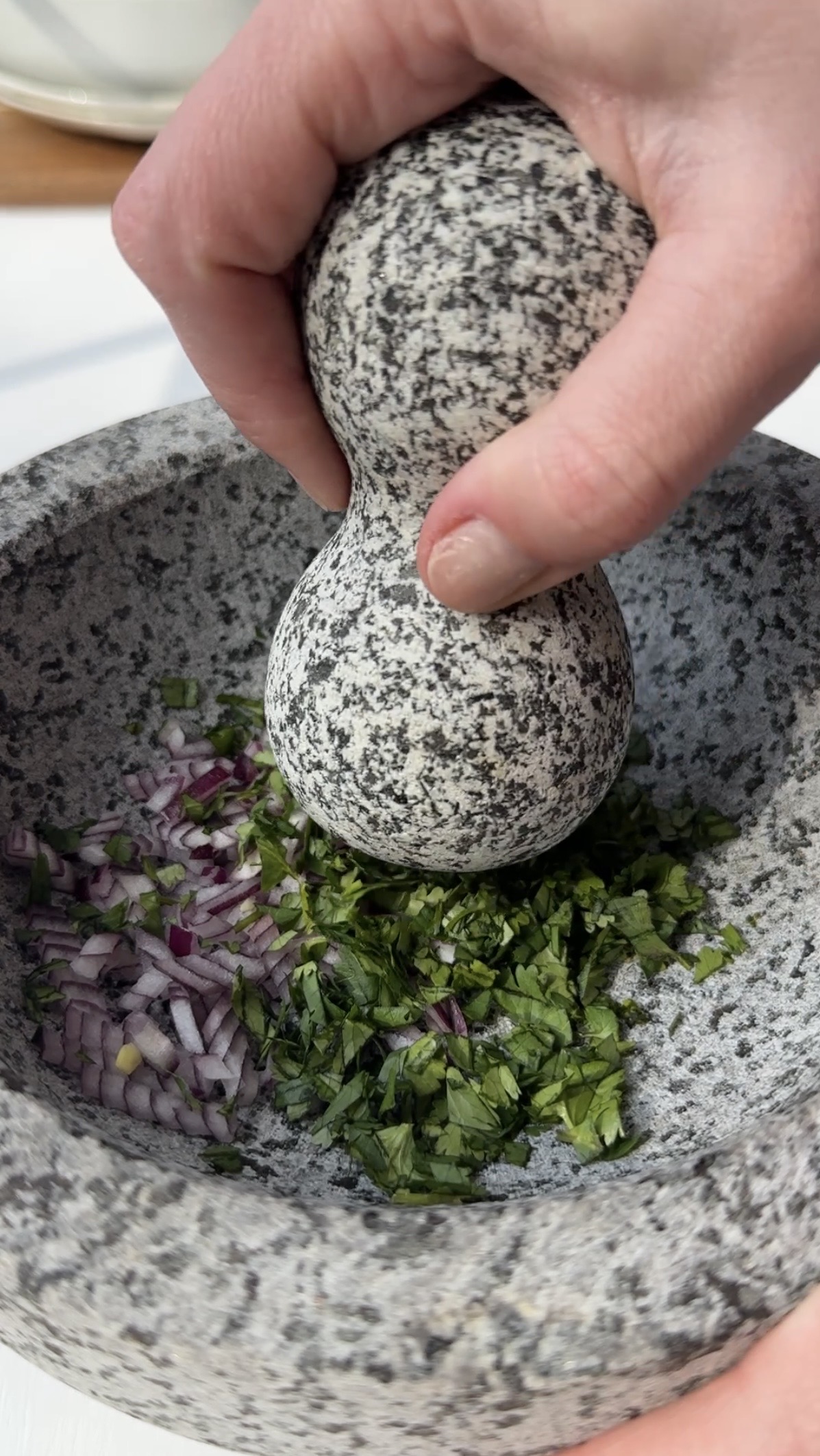 A hand uses a stone mortar and pestle to crush chopped red onions and fresh green herbs inside the bowl.
