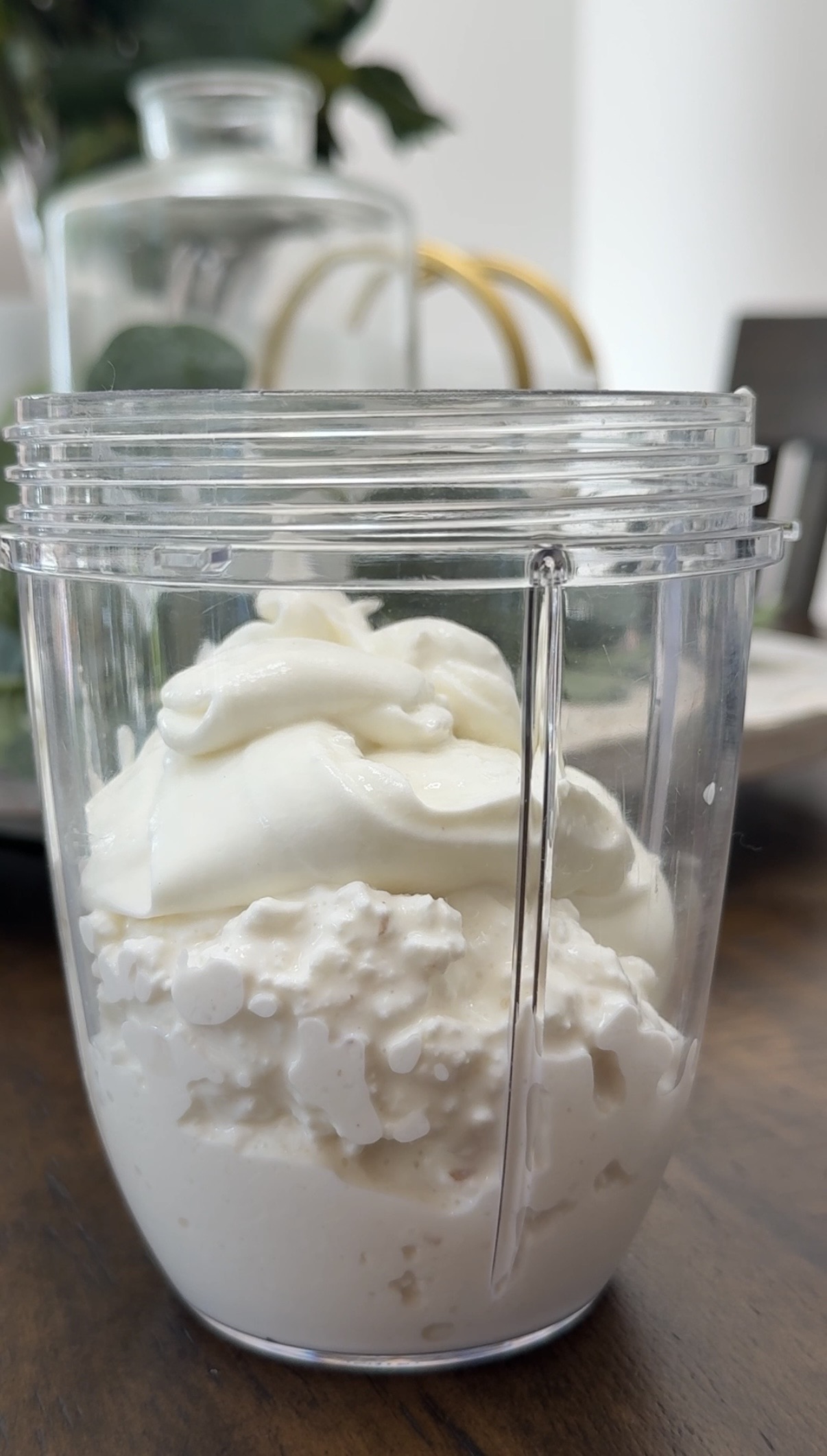 A clear blender cup filled with cottage cheese and a dollop of plain yogurt sits on a wooden table, with blurred household items in the background.