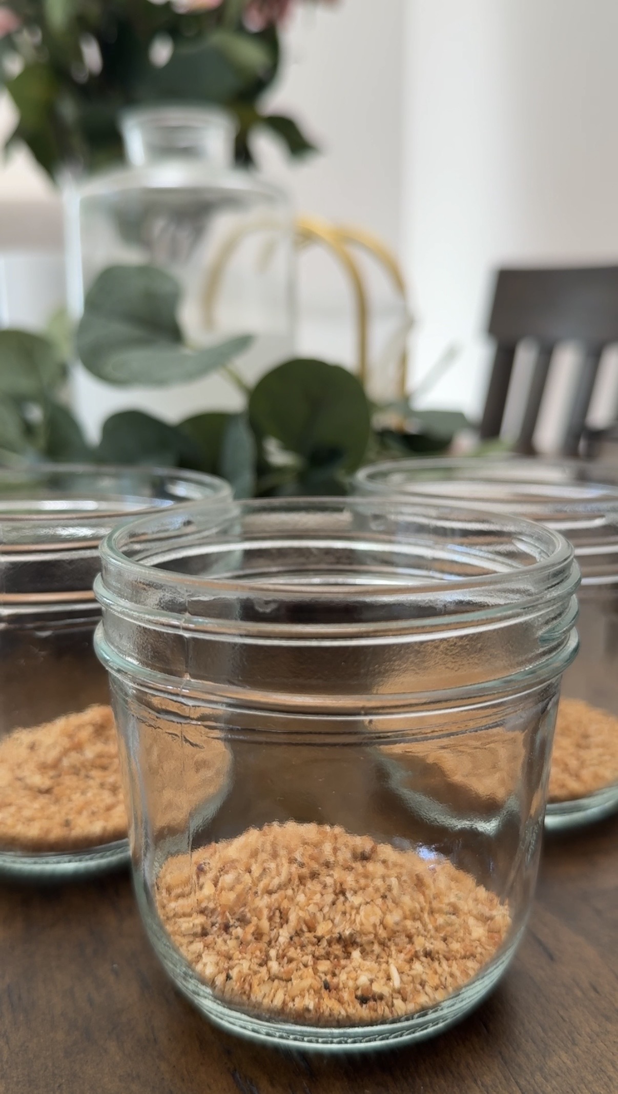 Three small glass jars, each containing a layer of brown crumbs, are arranged on a wooden table. Green foliage and a blurred water pitcher are visible in the background.