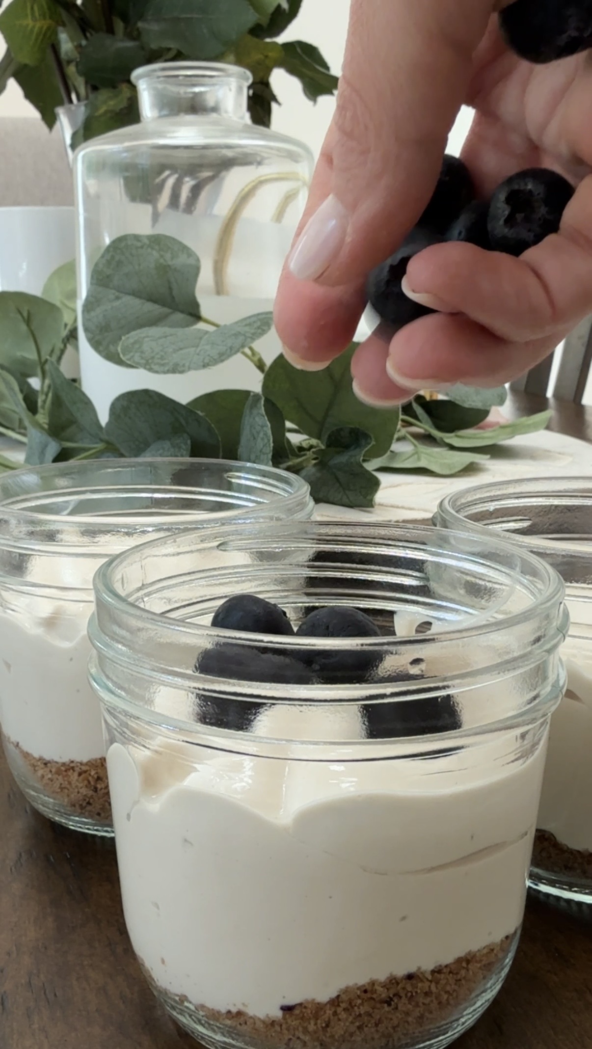 A hand places fresh blueberries on top of creamy dessert jars with a crumbly base. More dessert jars and green foliage are visible in the background.