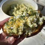 A hand holds a slice of toasted bread topped with creamy avocado, shredded hard-boiled egg, fresh dill, and lemon zest. In the background, there is a bowl of the same avocado and egg mixture.