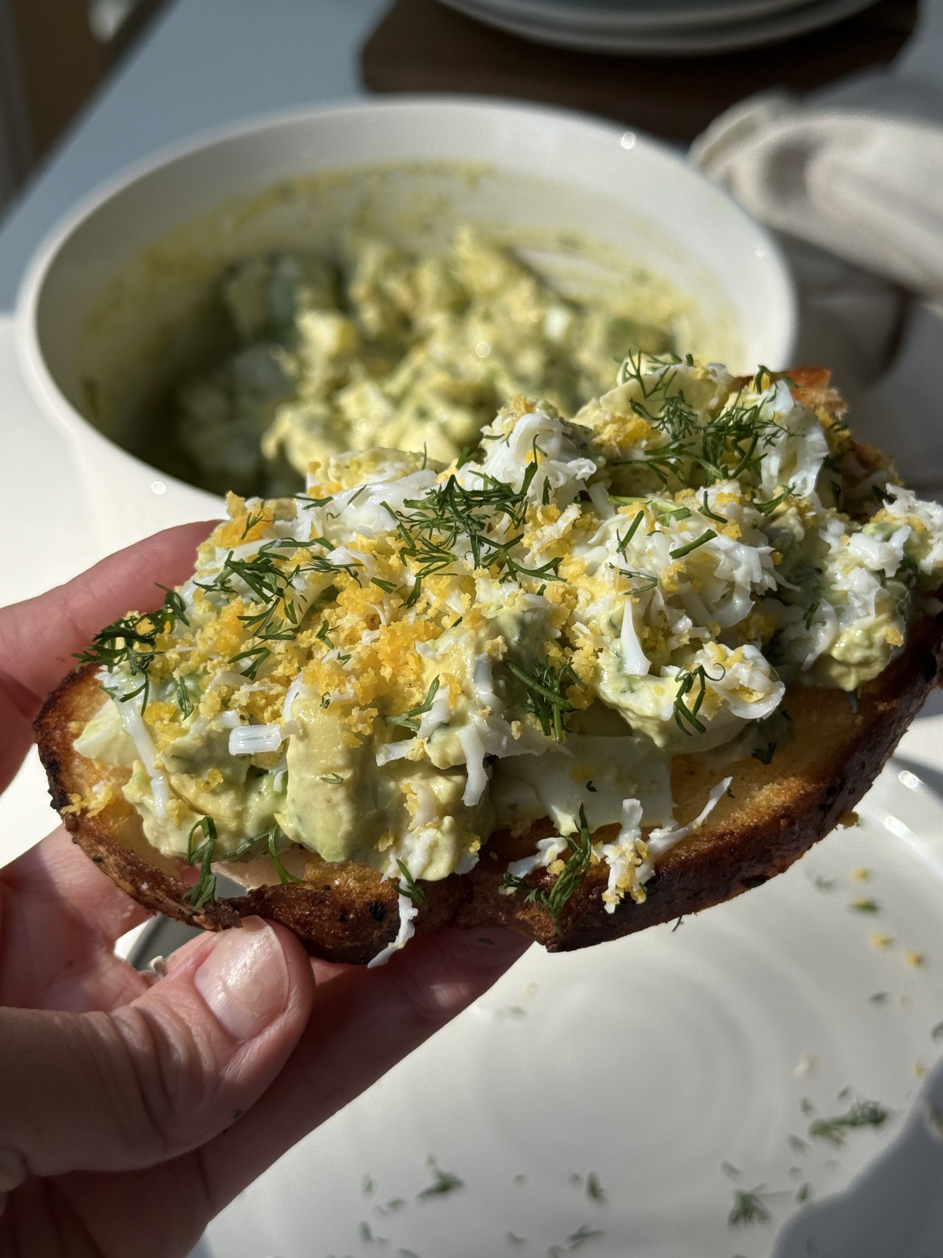 A hand holds a slice of toasted bread topped with creamy avocado, shredded hard-boiled egg, fresh dill, and lemon zest. In the background, there is a bowl of the same avocado and egg mixture.