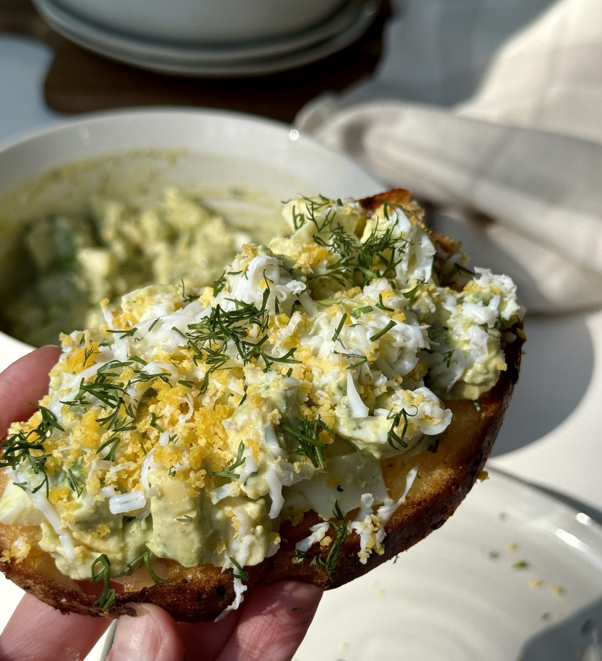 A hand holds a slice of toasted bread topped with avocado, grated boiled egg, fresh dill, and seasoning. A bowl with more avocado mixture and a napkin are visible in the background.