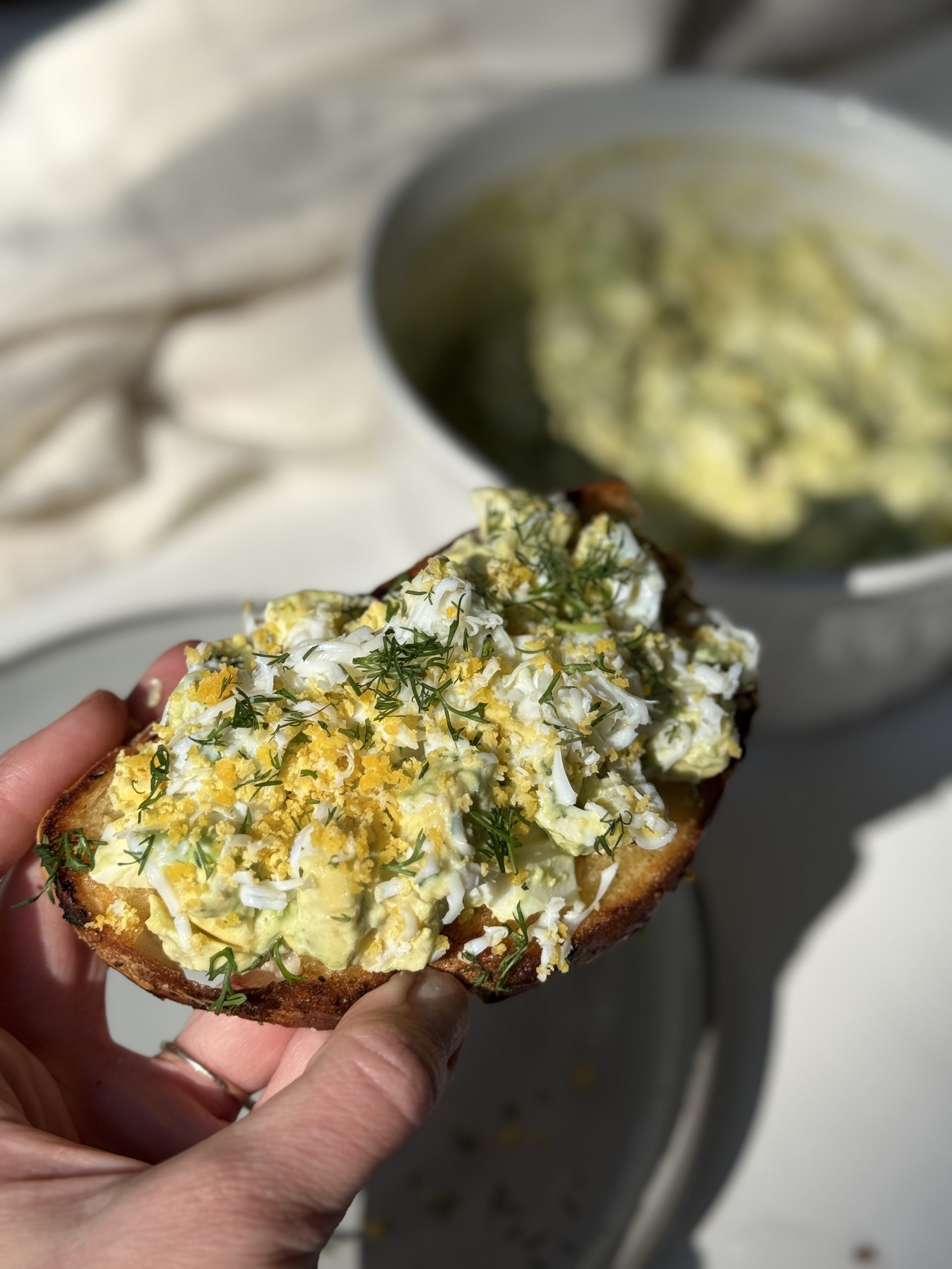 A hand holds a slice of toasted bread topped with a creamy egg salad and garnished with fresh dill. A bowl containing more egg salad is blurred in the background.