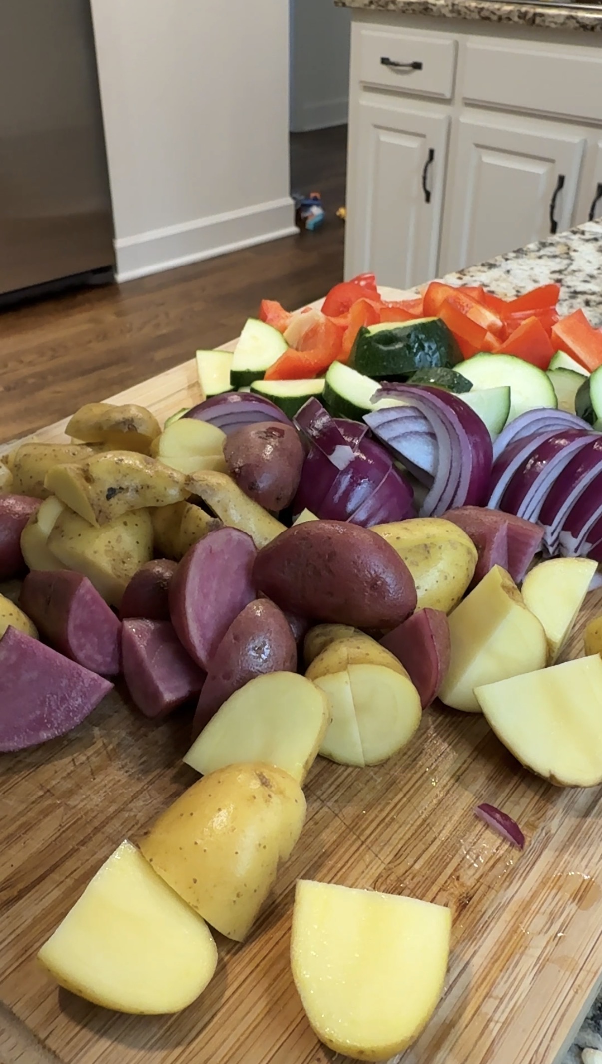 Wooden cutting board with chopped up potatoes, red onions, zucchini, and red bell peppers.