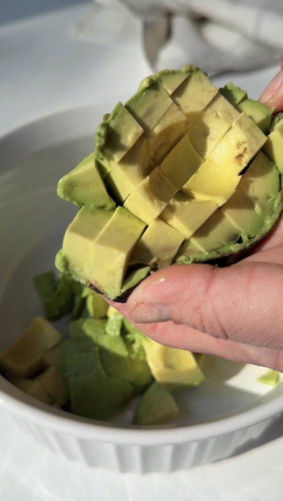 A hand holds half an avocado with its flesh cut into cubes, about to be scooped into a white bowl containing more avocado cubes.