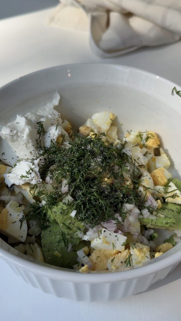 A white bowl filled with chopped hard-boiled eggs, avocado slices, onions, fresh herbs, and a dollop of soft white cheese, all mixed together on a white surface.
