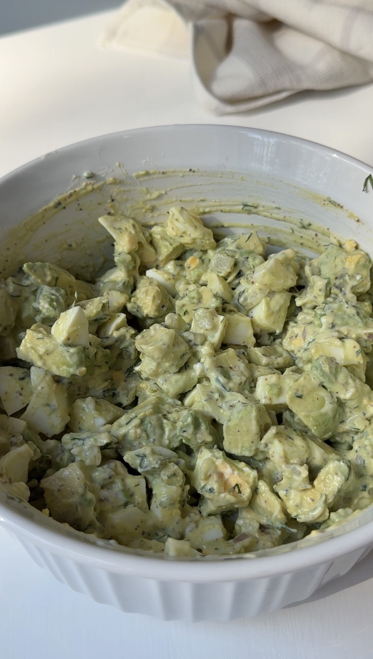 A white bowl filled with a creamy, chunky salad made of diced eggs and herbs mixed with a greenish dressing, possibly an egg salad with herbs.