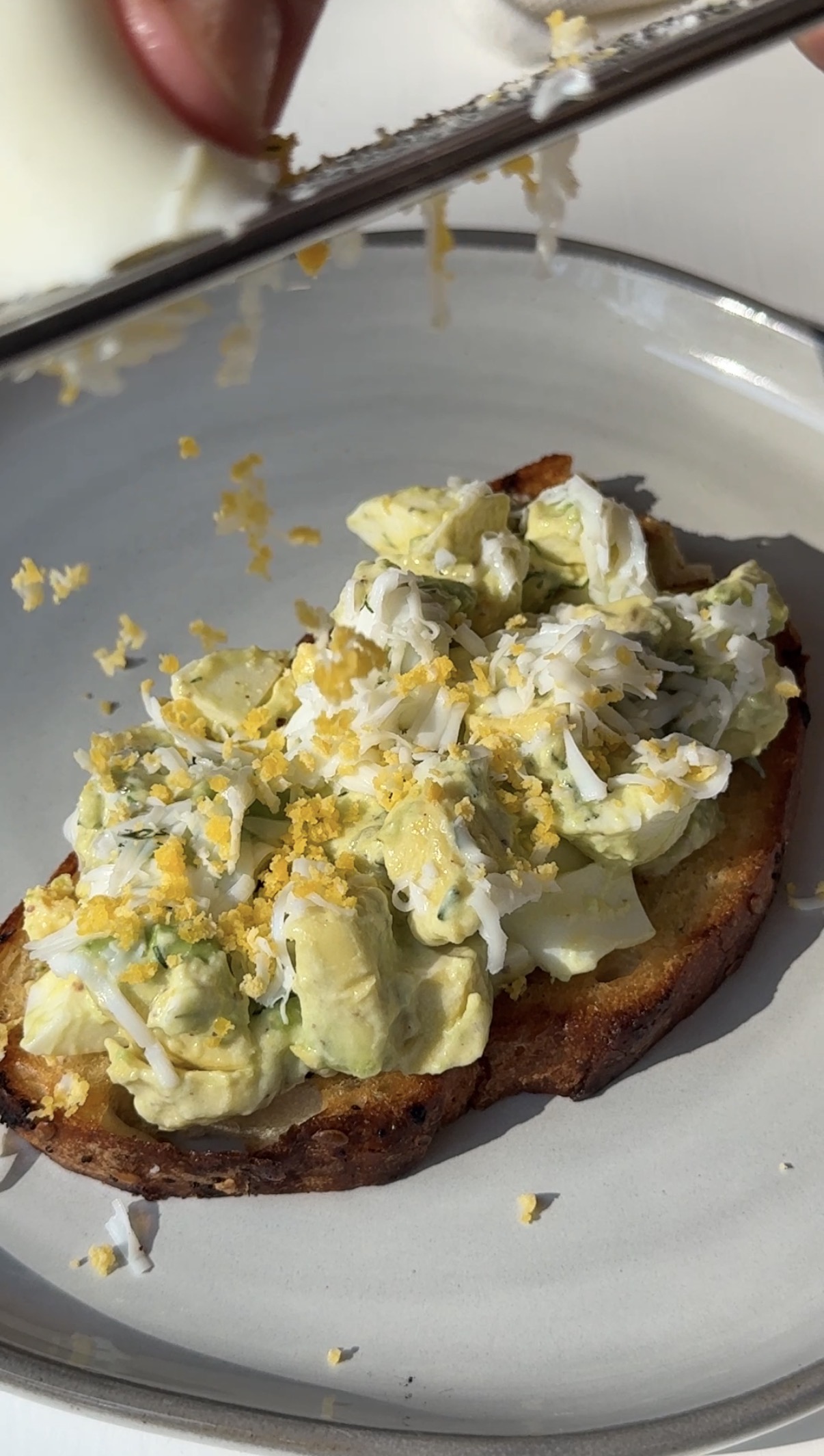 A slice of toasted bread topped with a creamy egg salad mixture and freshly grated hard-boiled egg, served on a white plate.