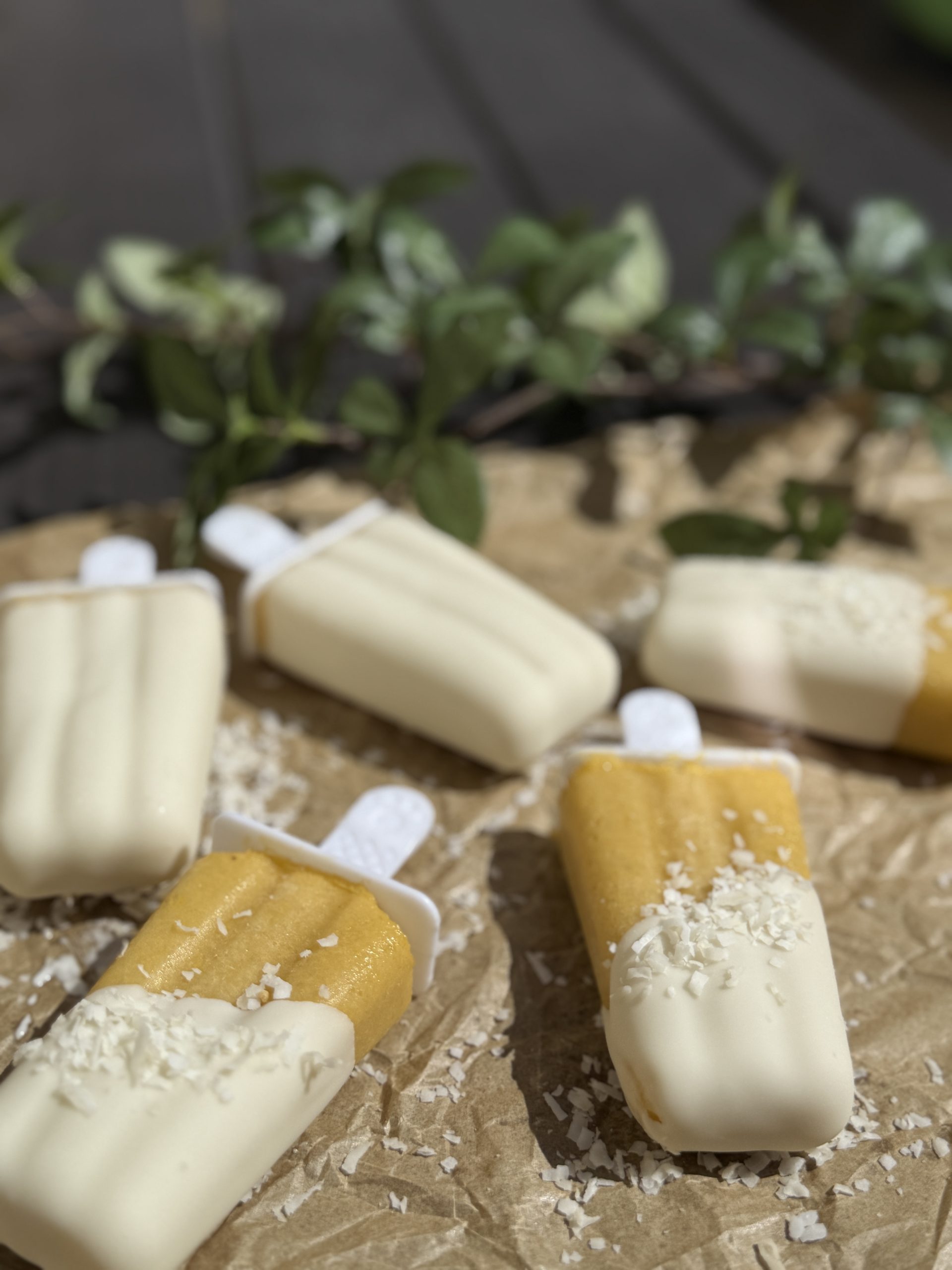 Five popsicles, partly coated in white chocolate and sprinkled with shredded coconut, are arranged on crumpled brown parchment paper with green leaves in the background.