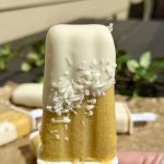 A hand holds a two-toned popsicle with a white top half and yellow bottom half, topped with shredded coconut, outdoors with more popsicles and greenery in the blurred background.