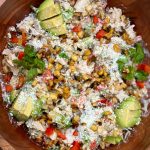 A wooden bowl filled with a mixed salad featuring creamy dressing, diced avocado, red bell peppers, fresh cilantro, roasted corn kernels, grated cheese, and shredded chicken.