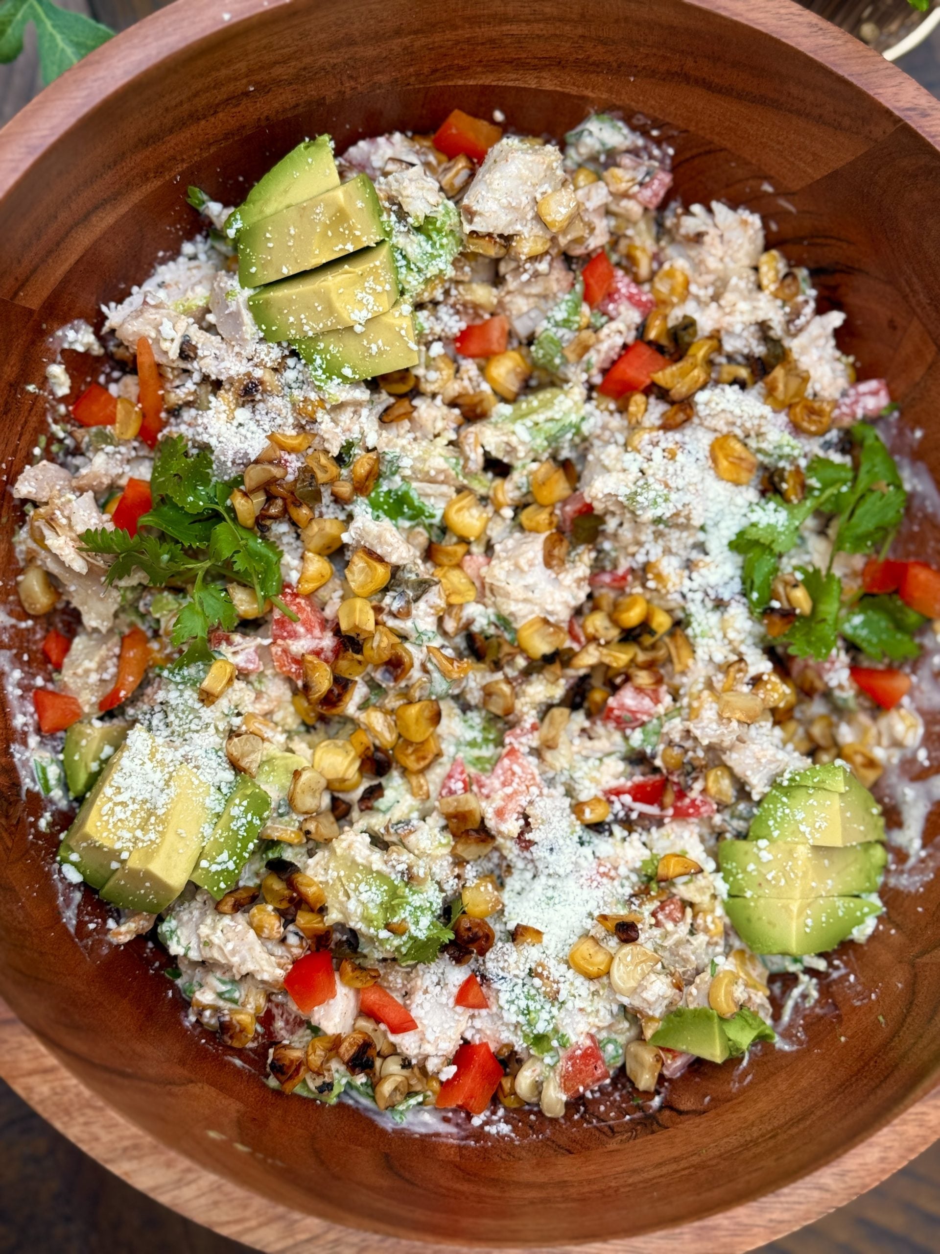 A wooden bowl filled with a mixed salad featuring creamy dressing, diced avocado, red bell peppers, fresh cilantro, roasted corn kernels, grated cheese, and shredded chicken.