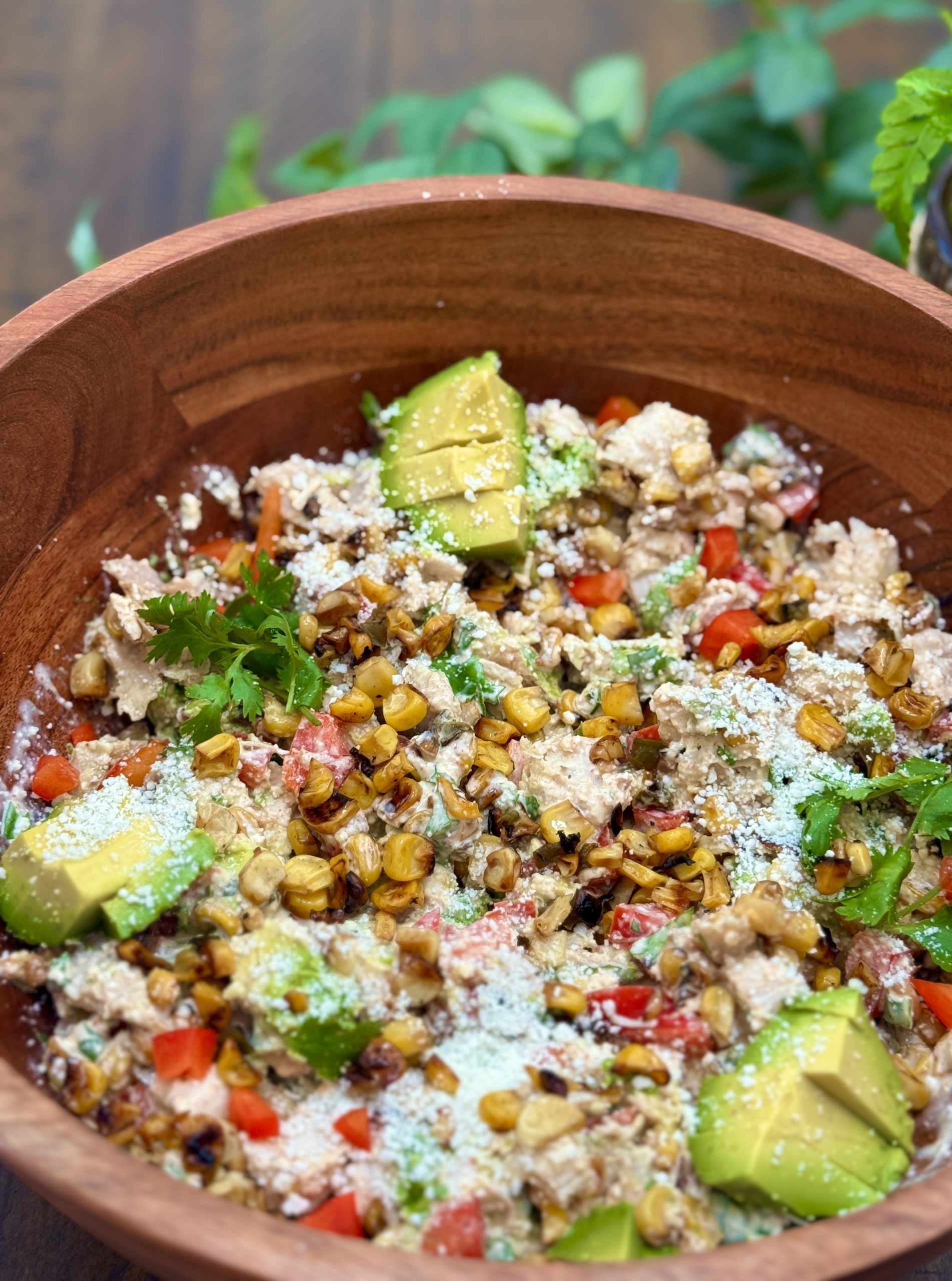 A wooden bowl filled with a colorful salad featuring grilled corn, diced tomatoes, avocado slices, fresh cilantro, crumbled cheese, and a creamy dressing. The background shows green leaves slightly out of focus.