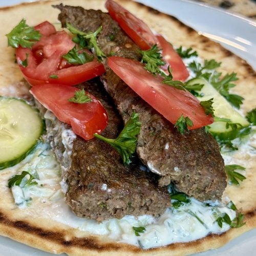 a plate with pita bread, tzatziki sauce and juicy oven-baked beef kebabs, topped with cucumber and tomatoes