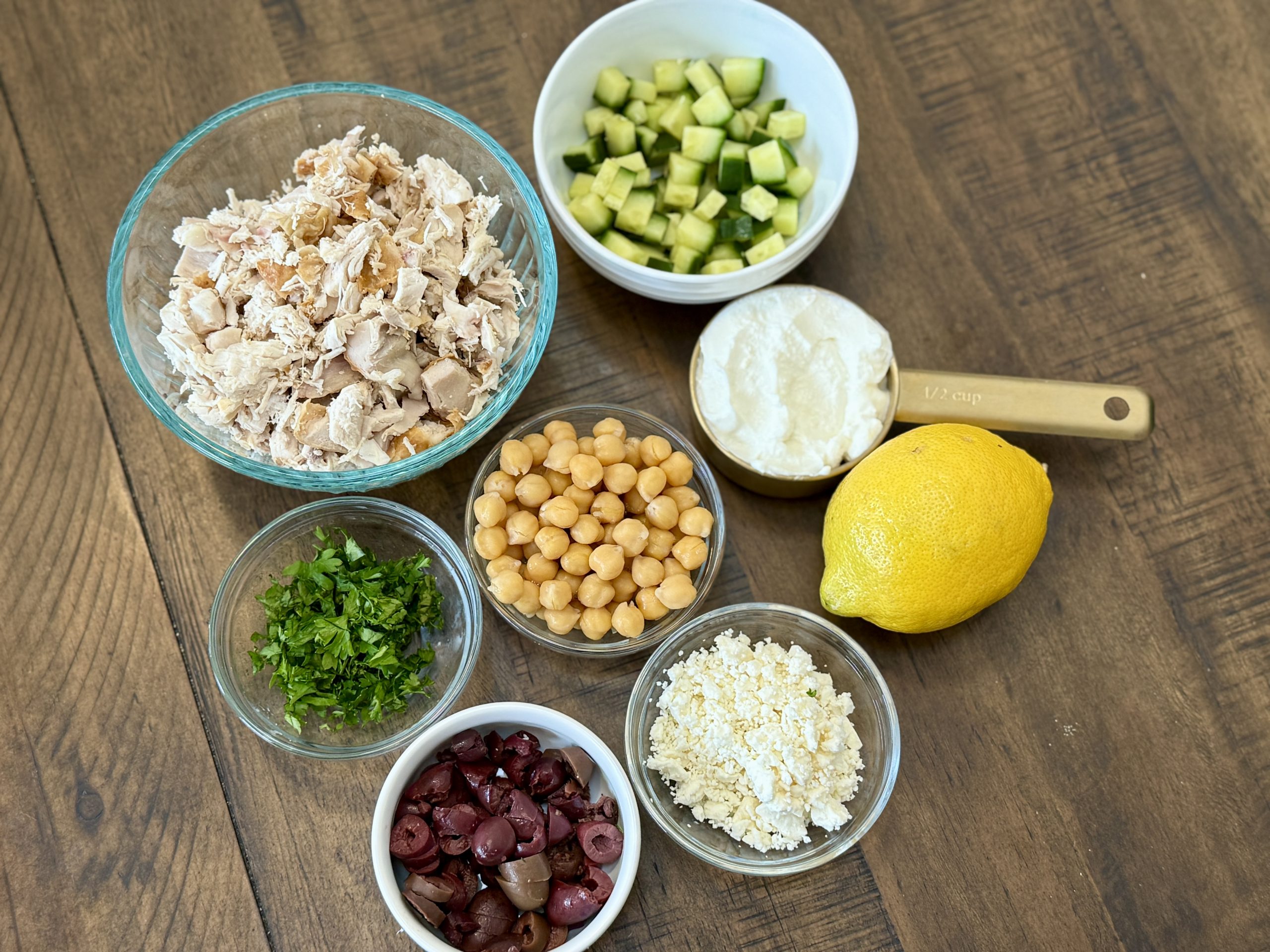 Seven bowls and a lemon on a wooden table hold chopped chicken, diced cucumber, Greek yogurt, chickpeas, fresh parsley, sliced Kalamata olives, and crumbled feta cheese.
